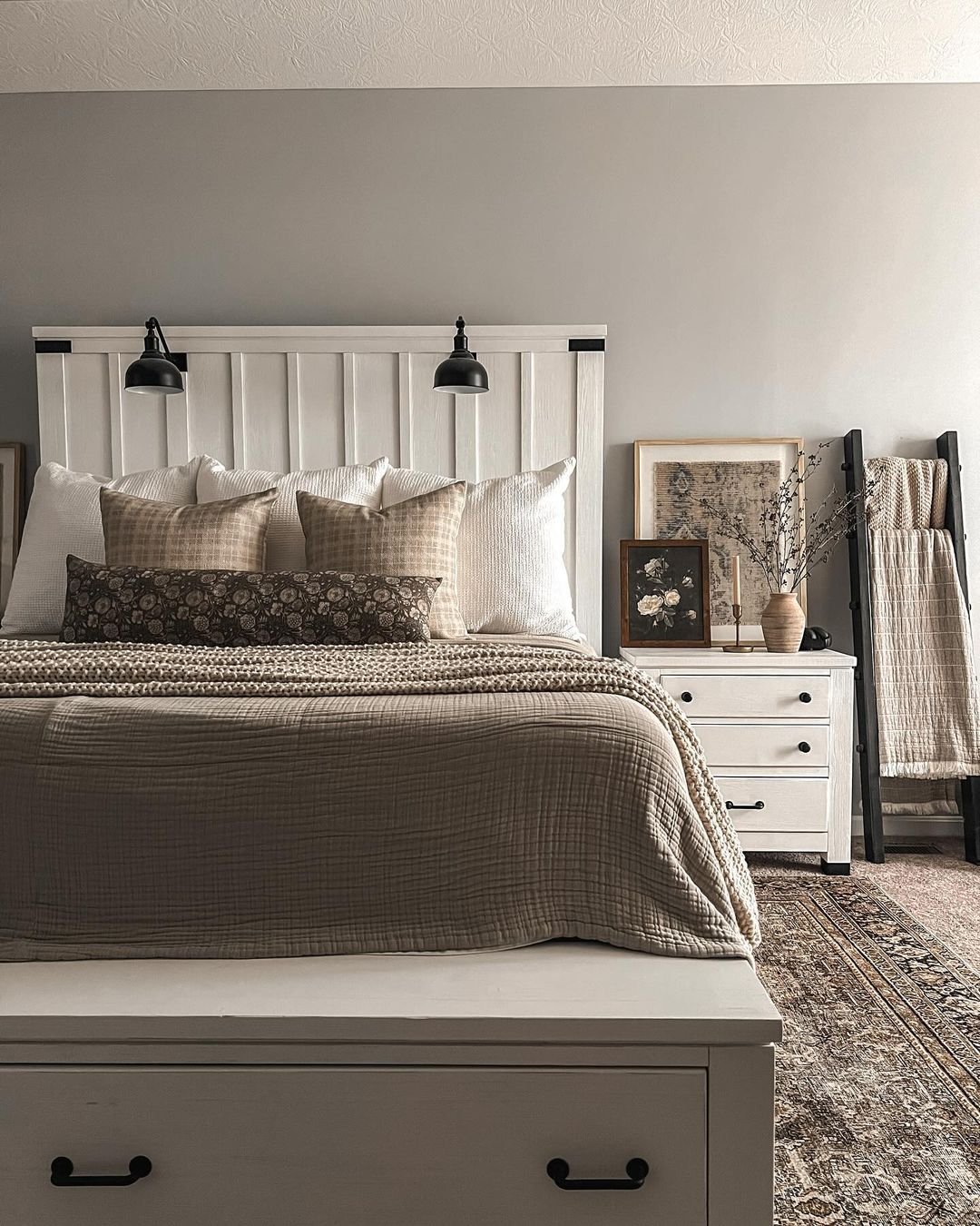 A cozy farmhouse bedroom with a white vertical shiplap headboard, a large beige quilt, an antique-style area rug, and a dark decorative ladder holding throw blankets.
