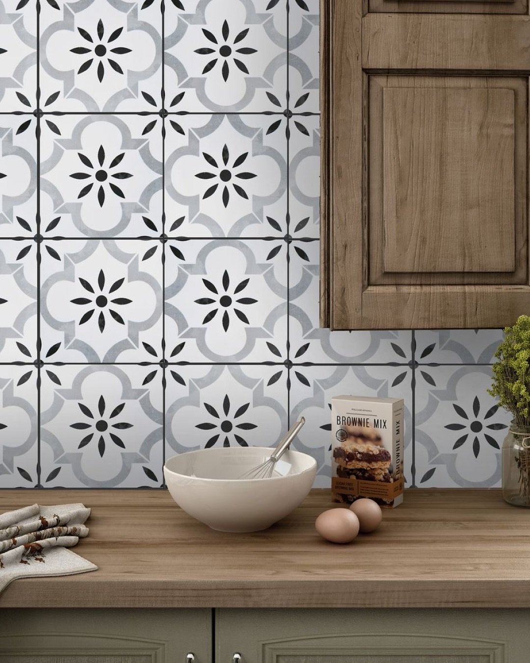 Kitchen backsplash featuring large square cement or porcelain tiles with a black, gray, and white repeating floral and quatrefoil pattern, paired with rustic wood cabinets.
