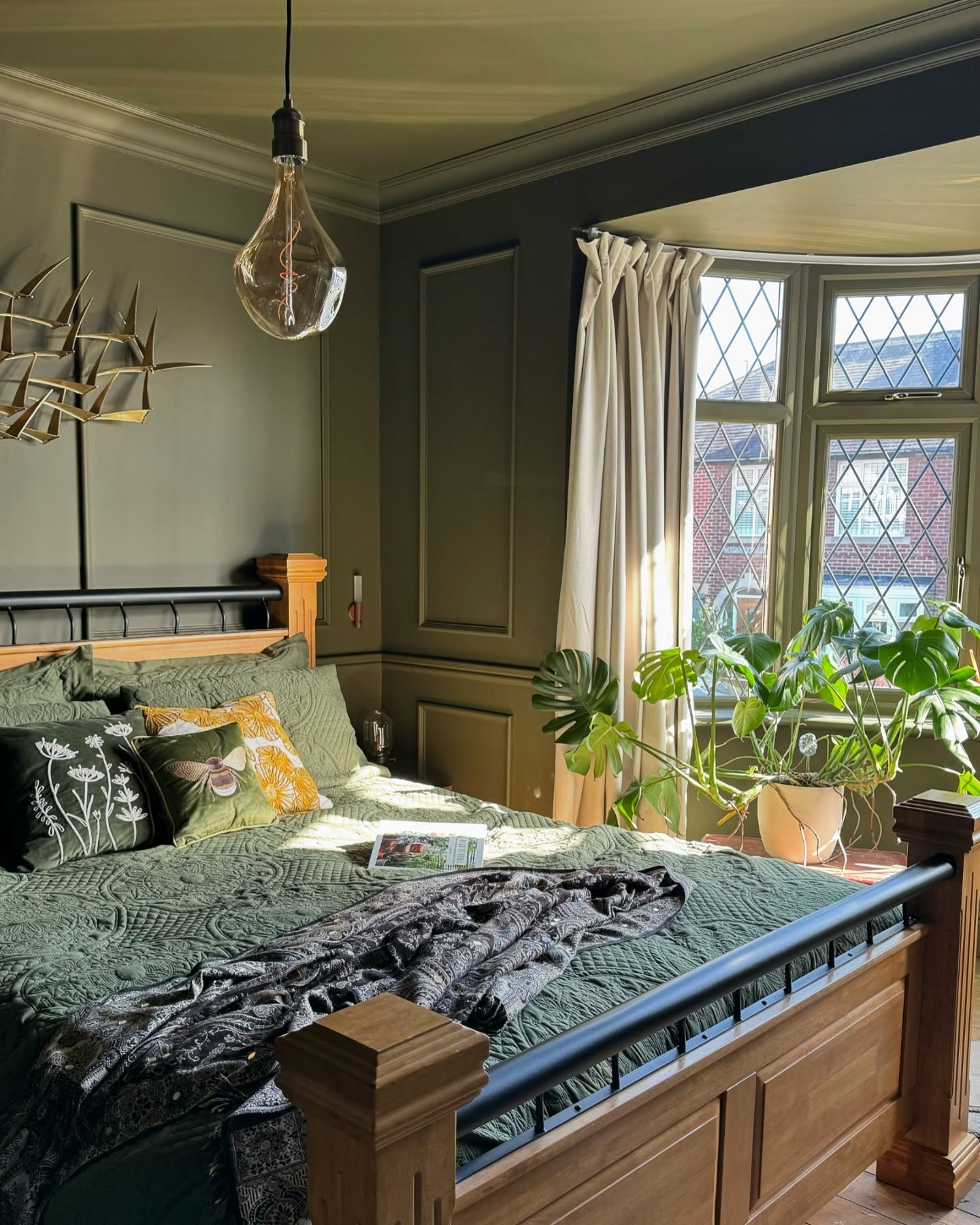 Dark olive green bedroom with wood paneling, a wooden four-poster bed, and a large Monstera plant in the sunlit bay window.