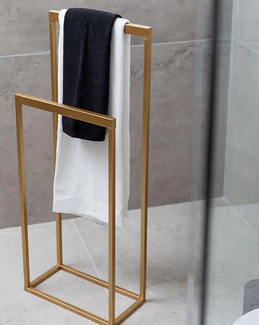 Freestanding, matte gold metal towel stand with two parallel vertical hanging bars, holding a folded black towel and a white towel.