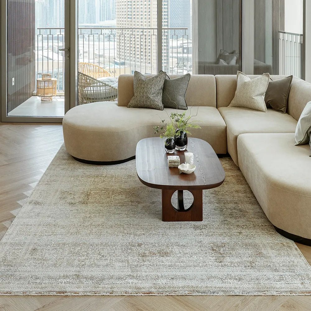 A modern beige living room with floor-to-ceiling windows overlooking a city skyline, featuring a curved sectional sofa, a dark wood coffee table, and a distressed area rug.