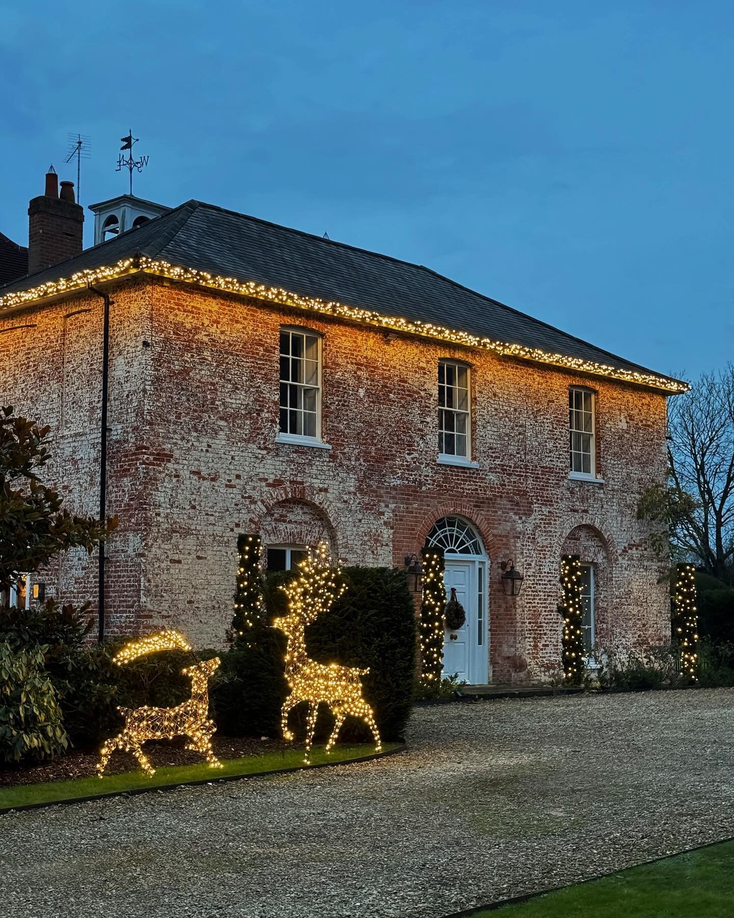 A historic brick home is outlined with warm white string lights along the roofline and windows, with two large, brightly lit wire-frame reindeer figures on the gravel drive.