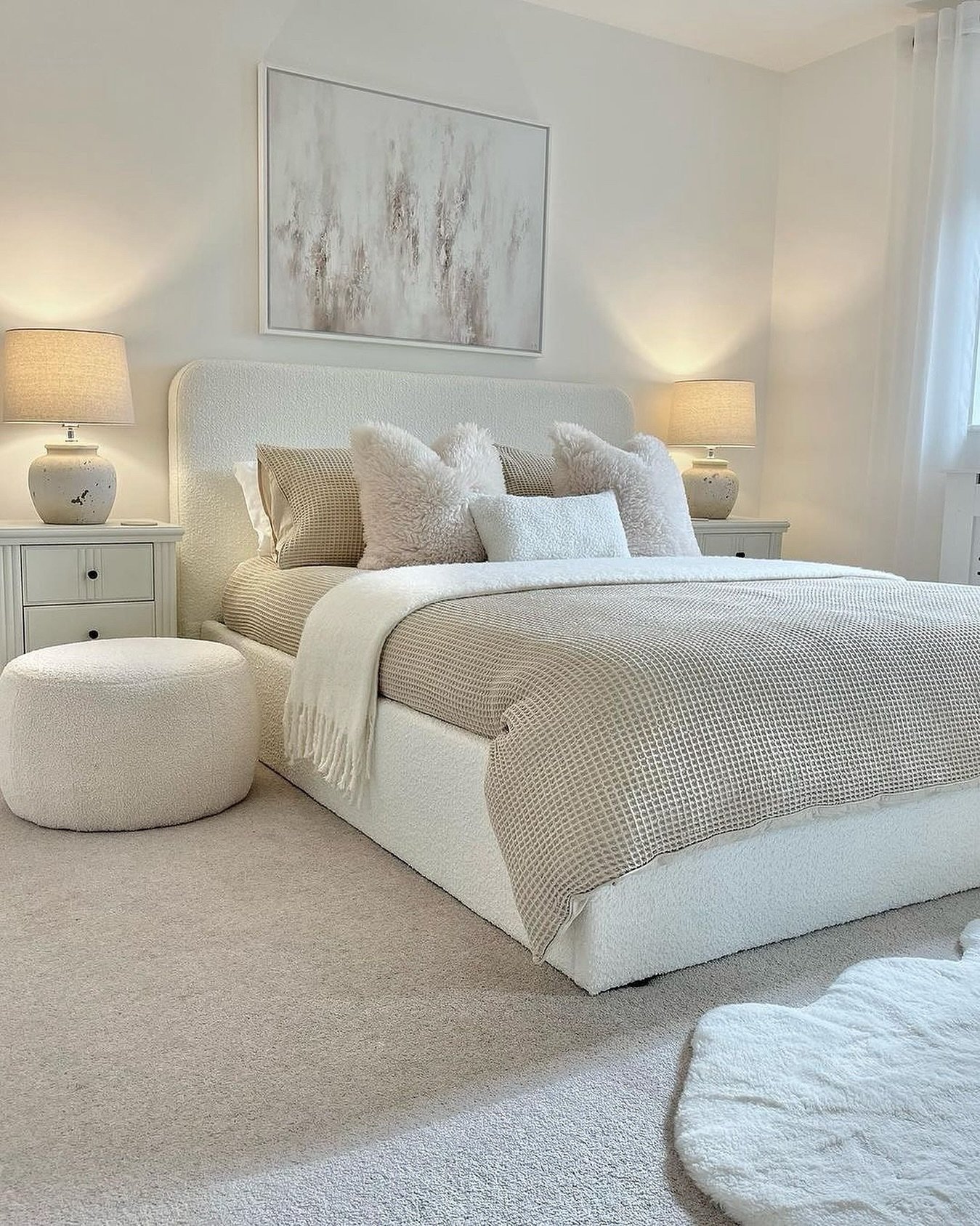 A cozy light beige bedroom featuring a bed and a round ottoman covered in soft bouclé fabric, a waffle knit blanket, and large abstract artwork over the headboard.