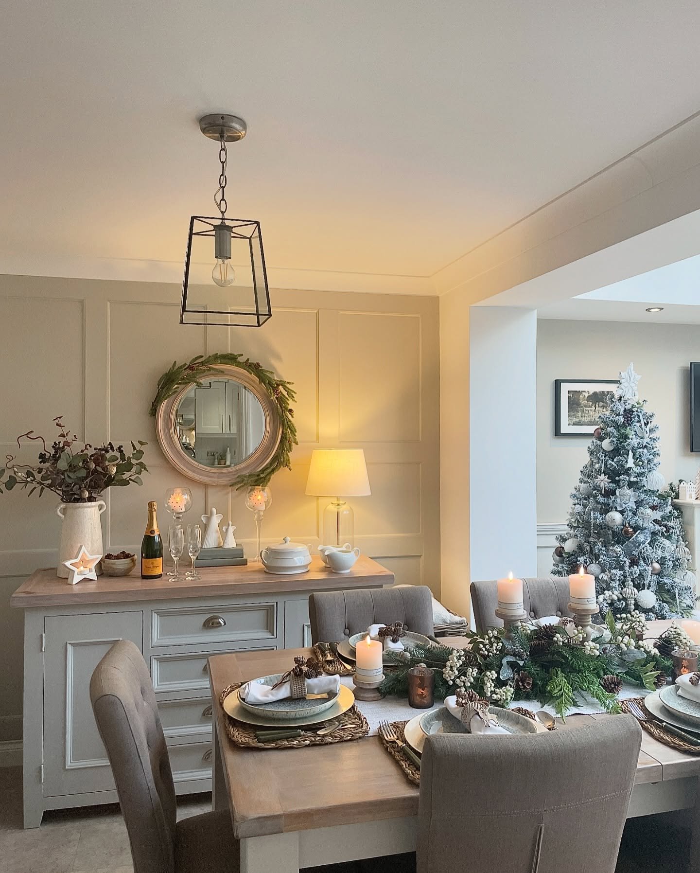 A dining table set with neutral upholstered chairs and a light wood surface, featuring woven placemats, layered white and sage green plates, a centerpiece of pine and eucalyptus, and soft candlelight.