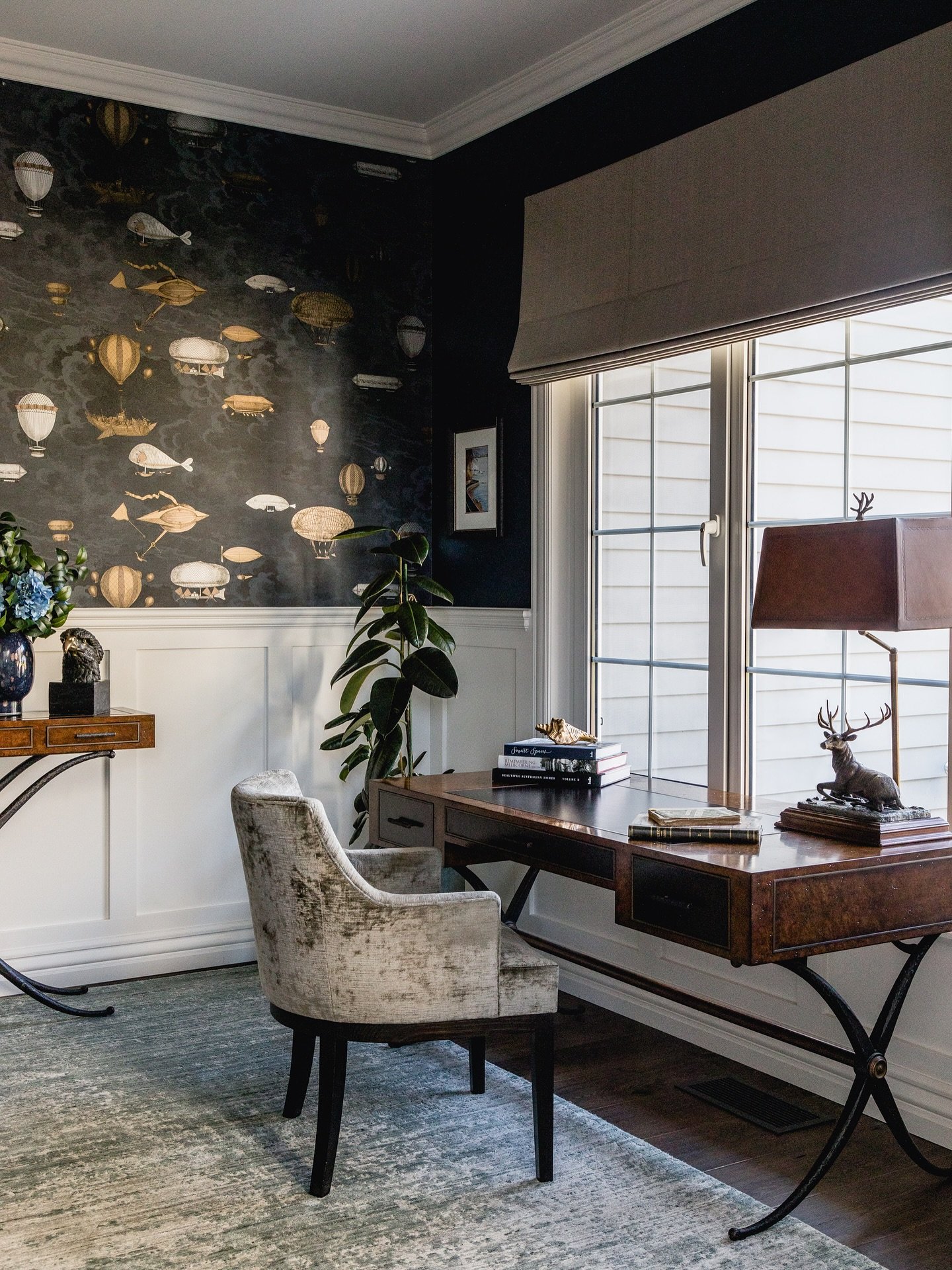 Dark, whimsical home office with hot air balloon wallpaper, white wainscoting, a burl wood desk, and a taupe velvet armchair.