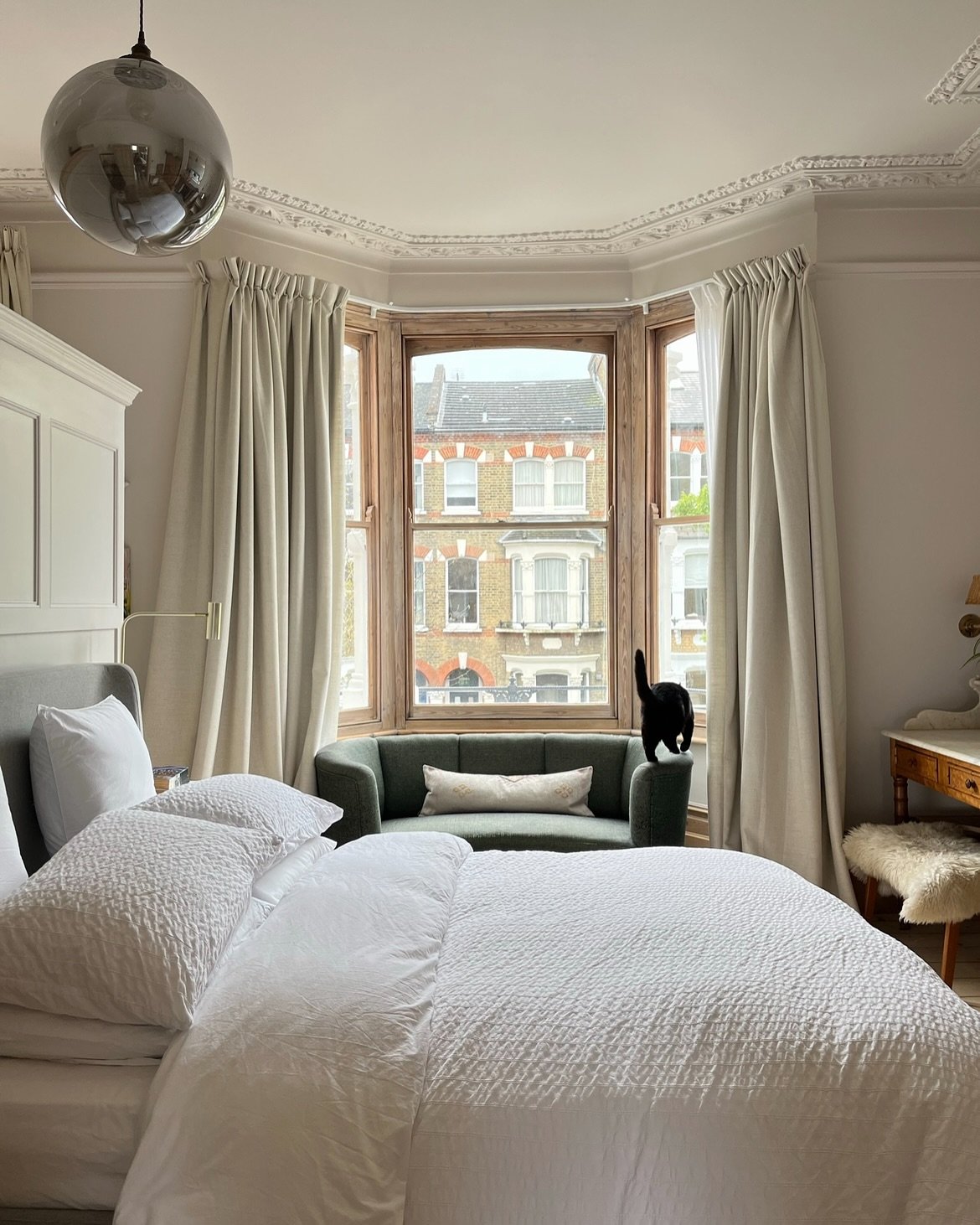 Cream linen curtains mounted on a track around a wooden-trimmed bay window in a bedroom.