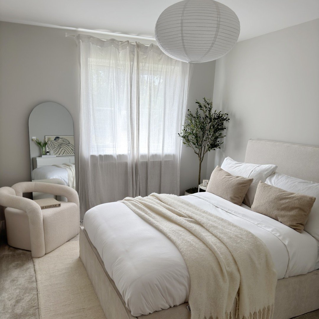 Small neutral bedroom featuring a plush fabric bed, a curved bouclé accent chair, a large paper lantern pendant, and a mirror leaning against the wall.