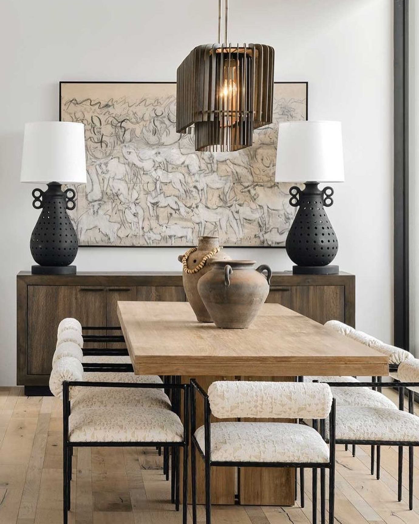 A dining room with a light wood table, boucle-style chairs with black metal frames, a dark wood sideboard, and a large abstract drawing flanked by black gourd-shaped lamps.