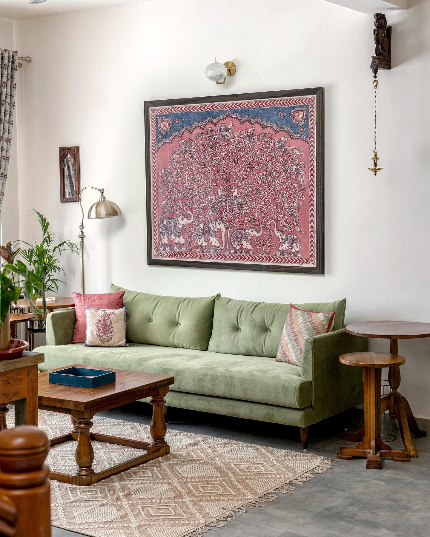 Large square framed Kalamkari or folk-style painting in red and blue depicting a Tree of Life with elephants, hung above a sage green sofa.