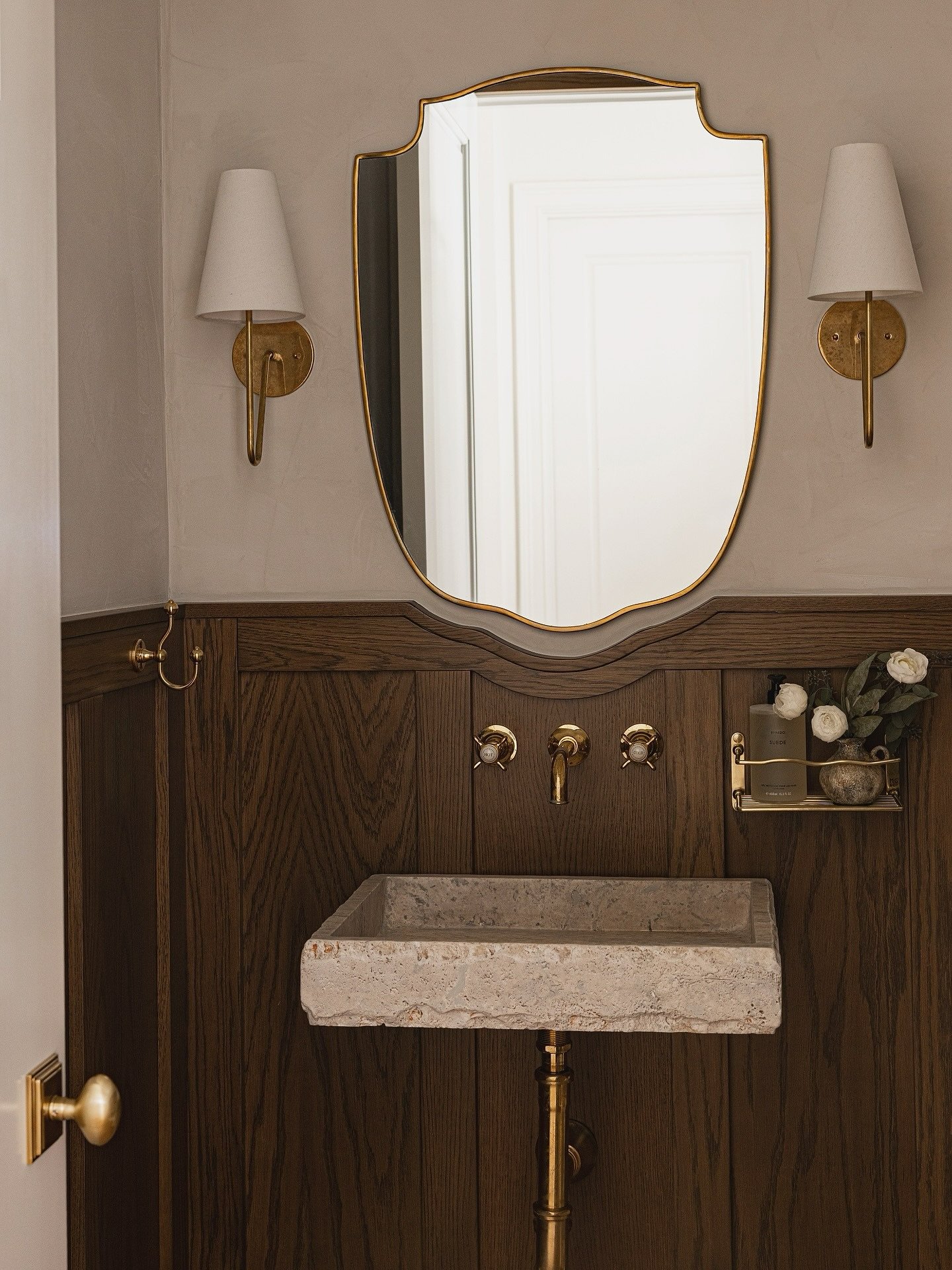 Wall-mounted stone trough sink over dark wood wainscoting, with a shapely mirror and brass sconces.