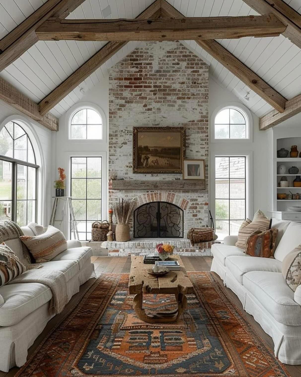 A bright, vaulted-ceiling living room featuring an exposed reclaimed wood A-frame, white slipcovered sofas, a whitewashed brick fireplace, and a distressed orange and blue vintage rug.