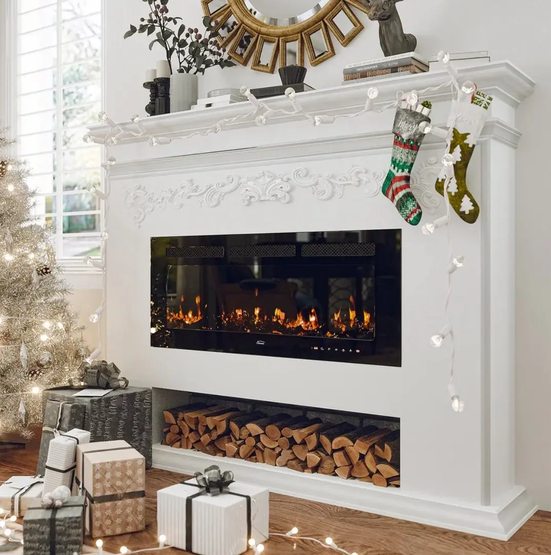 White, ornate electric fireplace mantel with carved detailing, a wide black firebox, and an open lower compartment filled with decorative logs, decorated for Christmas.