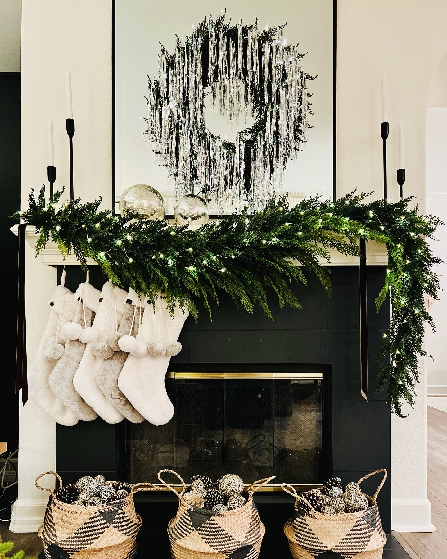Black tile fireplace with a simple, dark evergreen garland, five fuzzy white stockings with pom-poms, and a statement silver tinsel wreath hanging above.