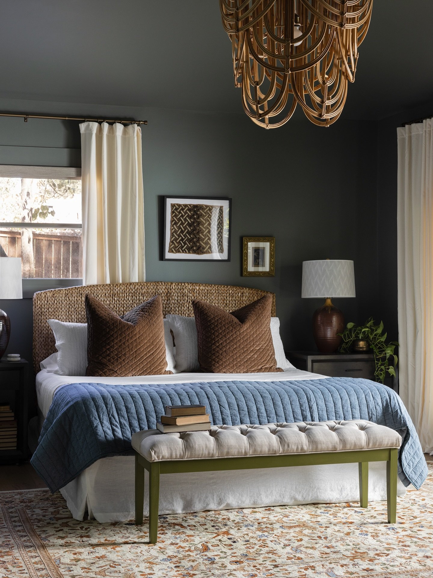A cozy, dark green bedroom with a rattan headboard and a button-tufted bench with light upholstery and dark green painted legs at the foot of the bed.