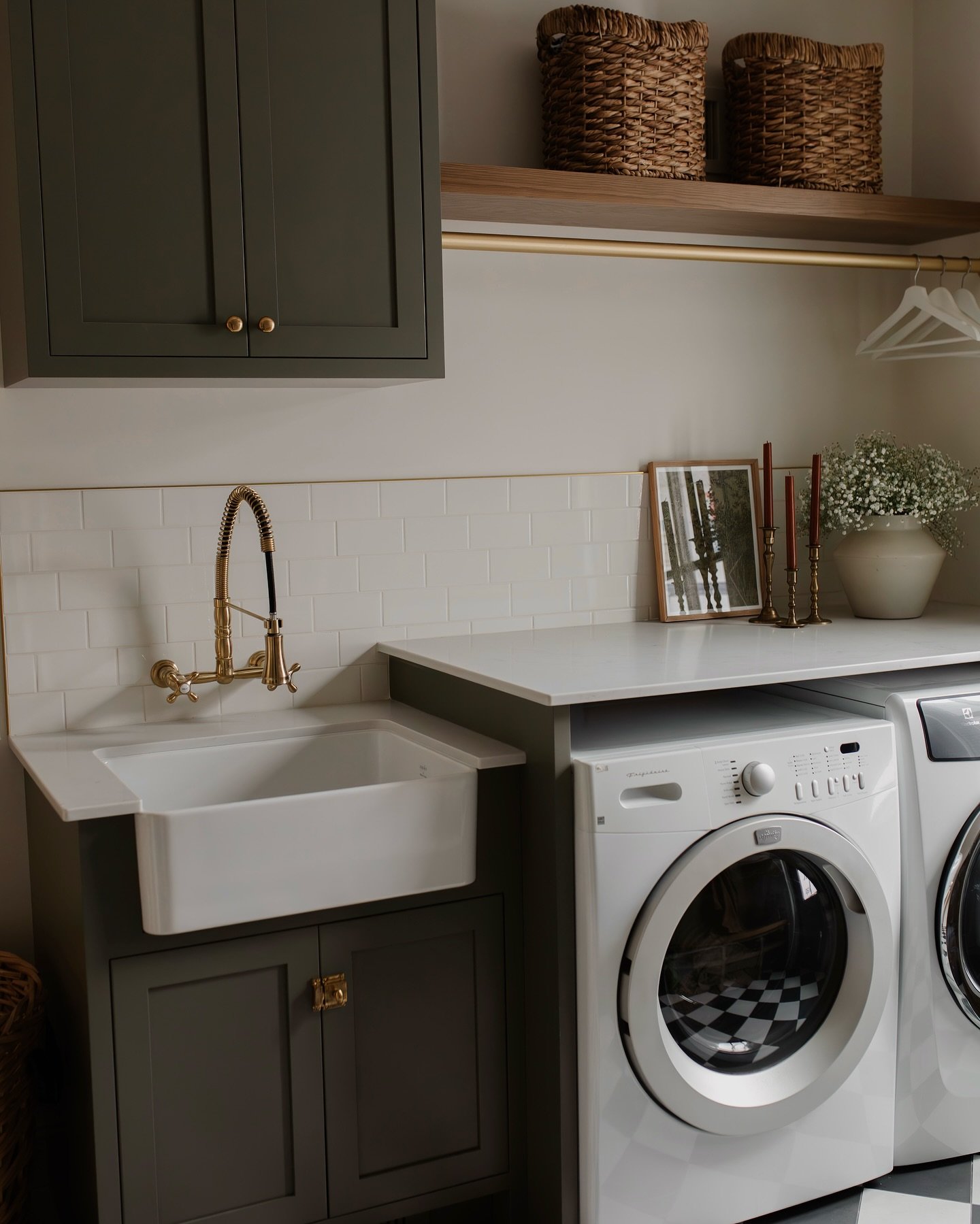 A small, luxury laundry room featuring a deep olive/khaki green cabinet with a white farmhouse sink, white subway tile backsplash trimmed with gold, and a wood shelf with a brass drying rod underneath.