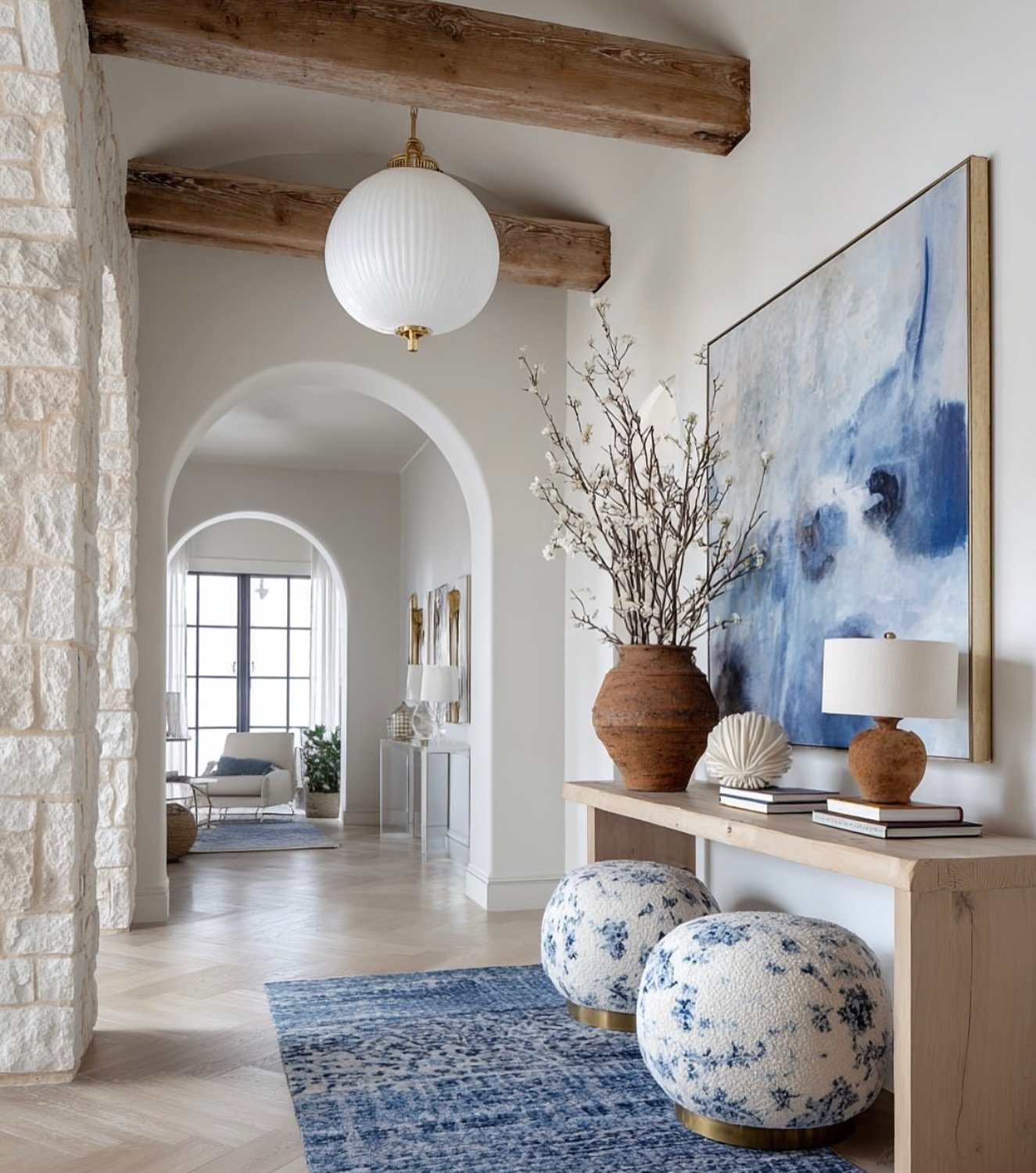 A wide hallway with exposed wood beams, a light wood console table, a large blue and white abstract painting, and two blue patterned poufs.