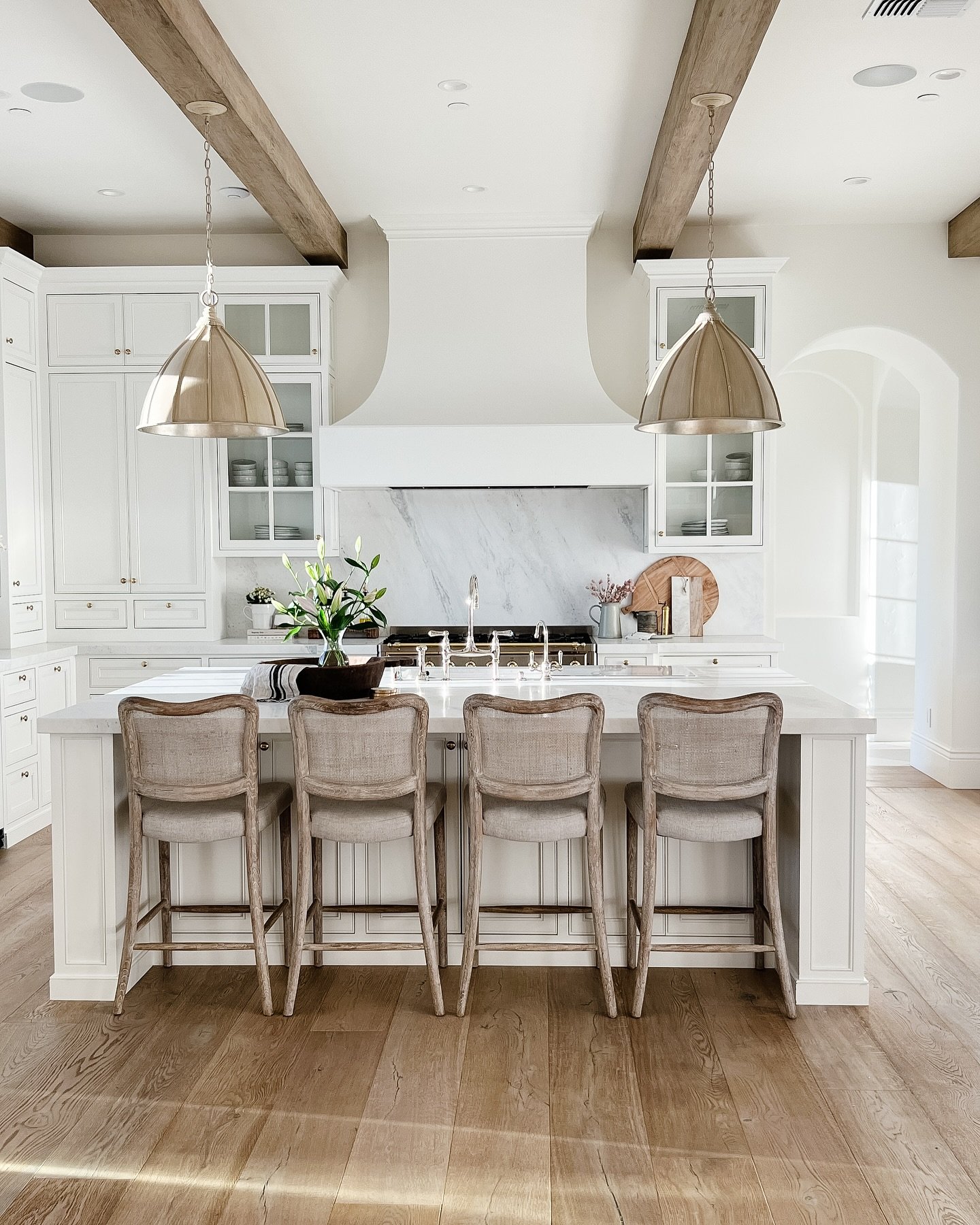 Elegant farmhouse kitchen with white cabinets, a full marble slab backsplash, light wood floors, rustic wood beams, and gold dome pendant lights over a white island.