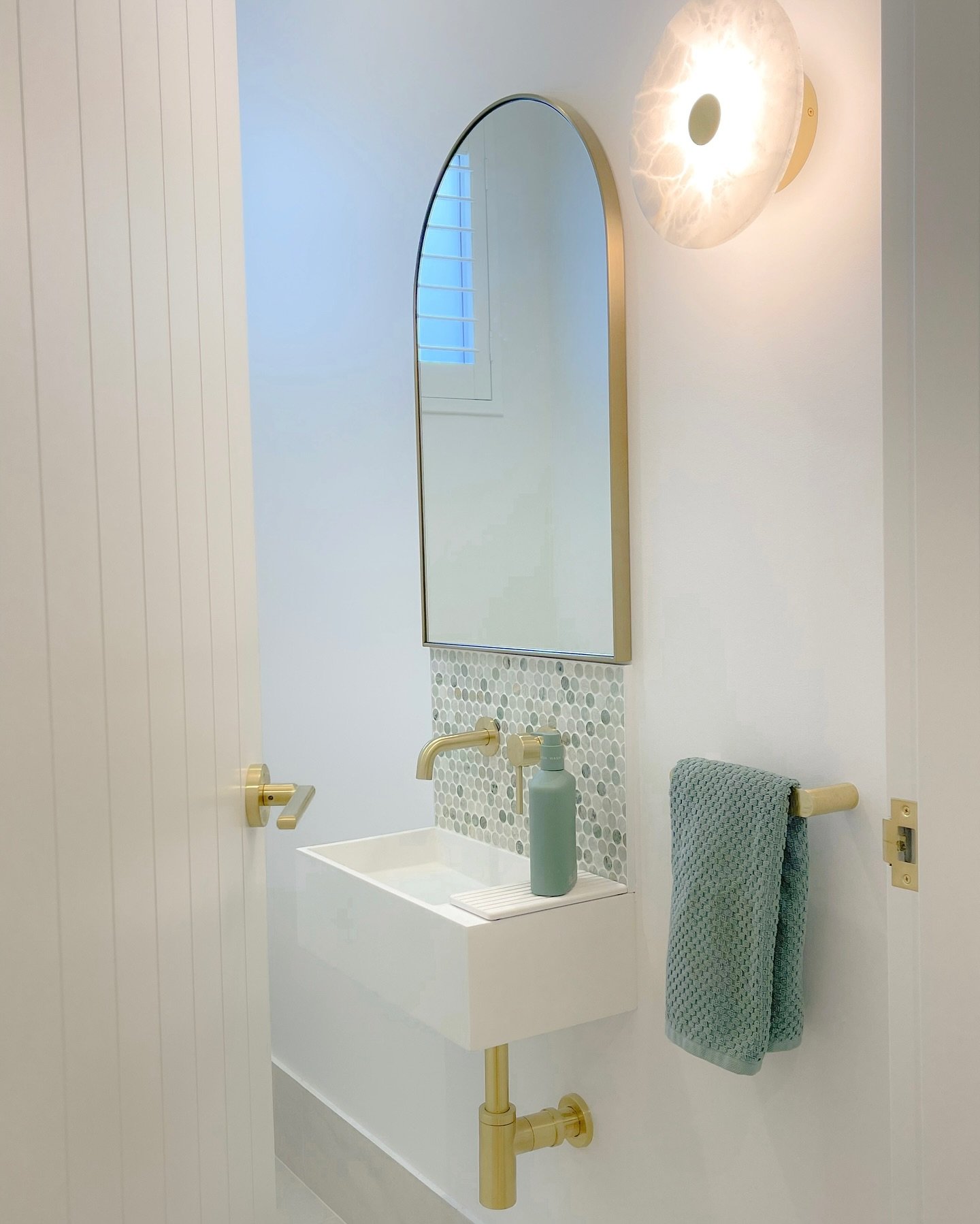 A short, horizontal brass hand towel bar mounted on a white wall between a sink and a door frame, holding a seafoam green towel.