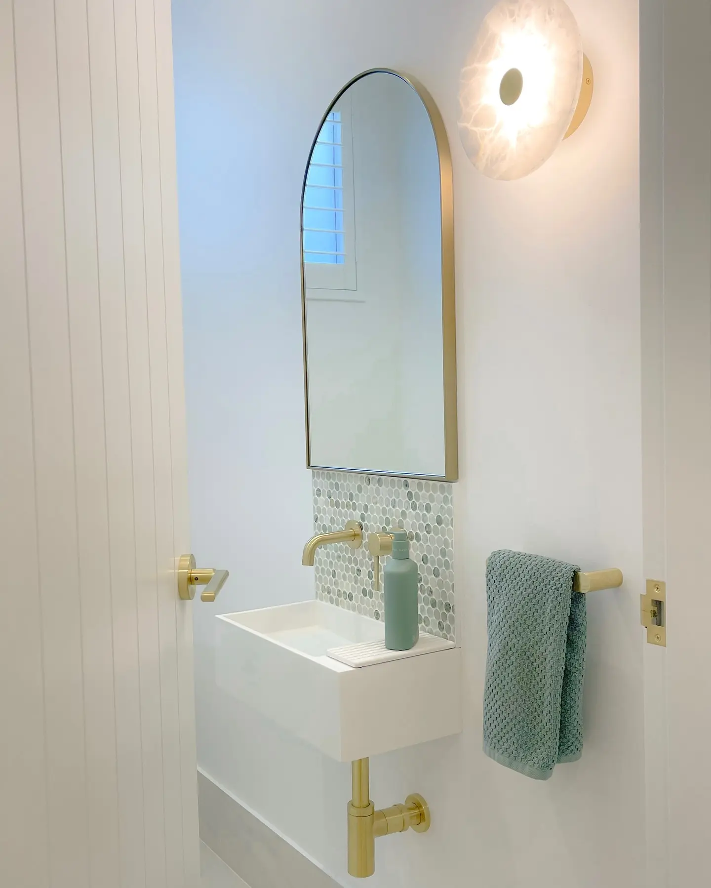 A short, horizontal brass hand towel bar mounted on a white wall between a sink and a door frame, holding a seafoam green towel.