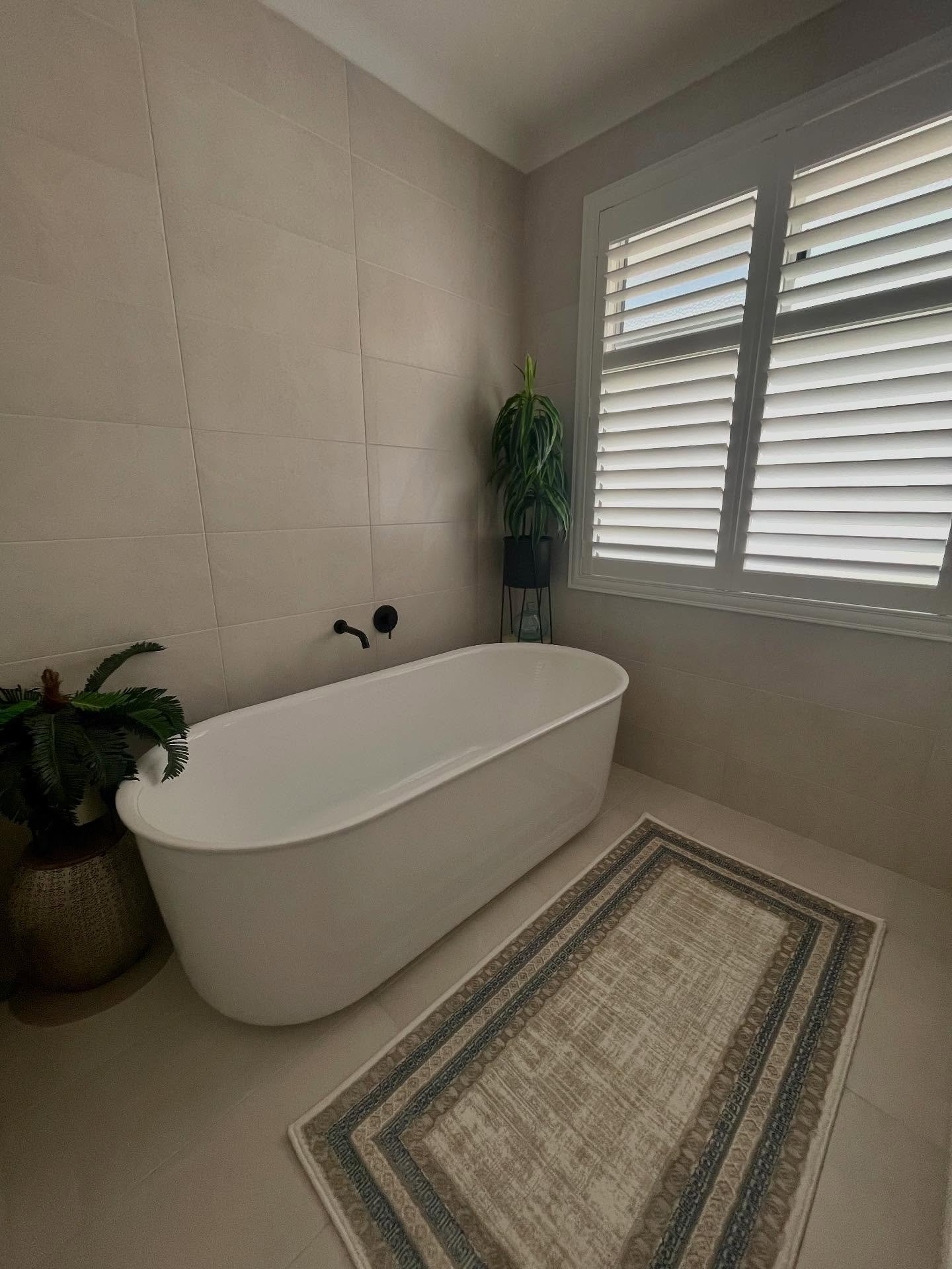 A contemporary bathroom featuring large, light beige tiles, a white freestanding tub, black wall-mounted fixtures, and a rectangular neutral rug with a detailed blue, gray, and brown geometric border pattern.