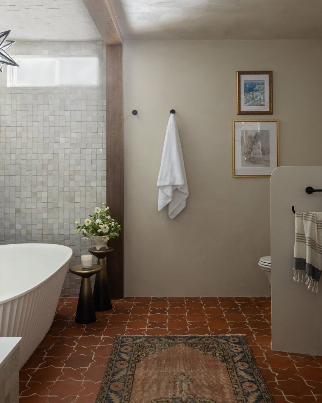 A warm, inviting bathroom with a standalone fluted tub, a creamy beige wall, and a floor covered in square terracotta tiles laid in a diagonal pattern.