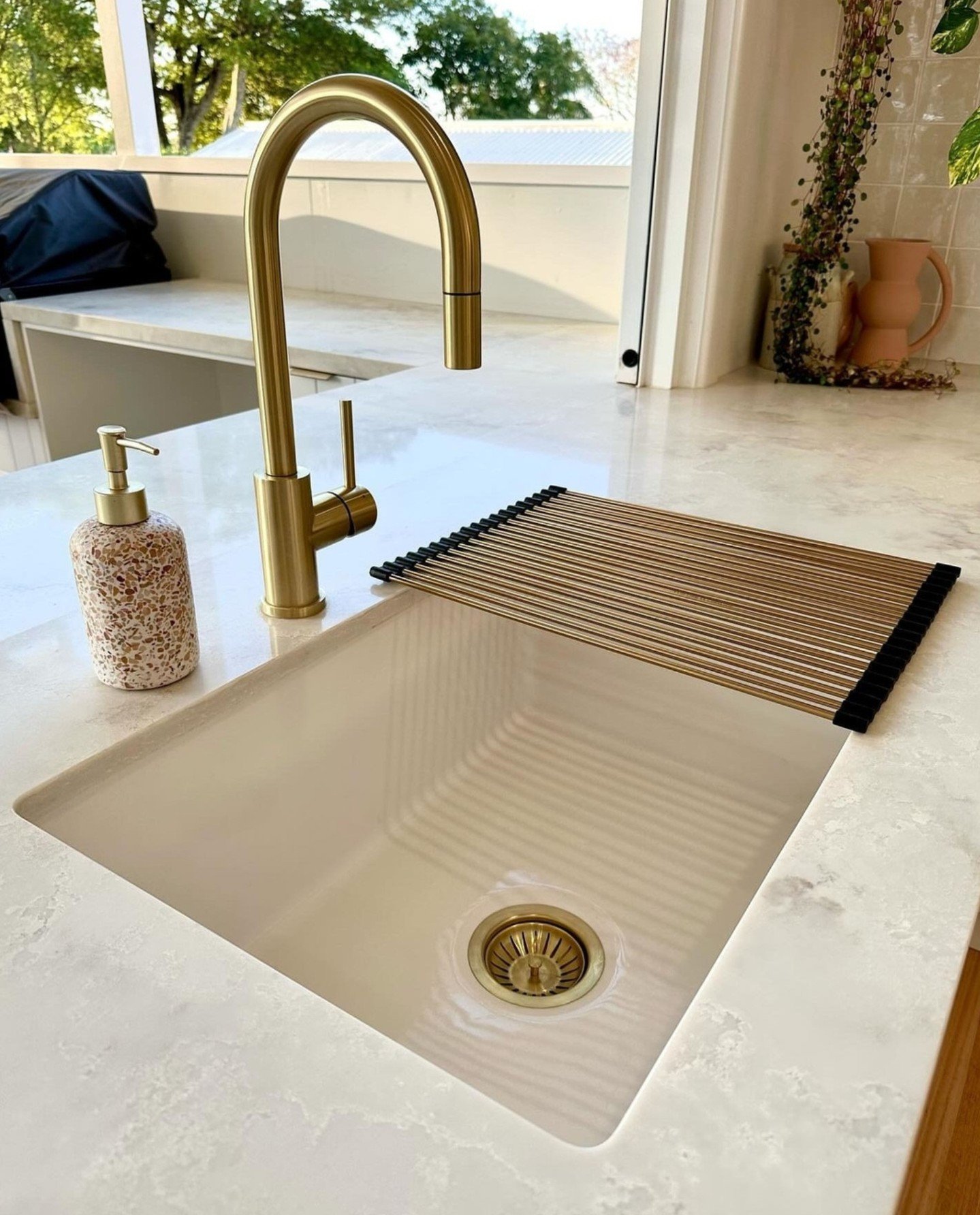 A close-up of a smooth, single-bowl beige or cream-colored undermount kitchen sink set in a light quartz countertop, featuring a curved brushed gold faucet and a gold roll-up drying rack.