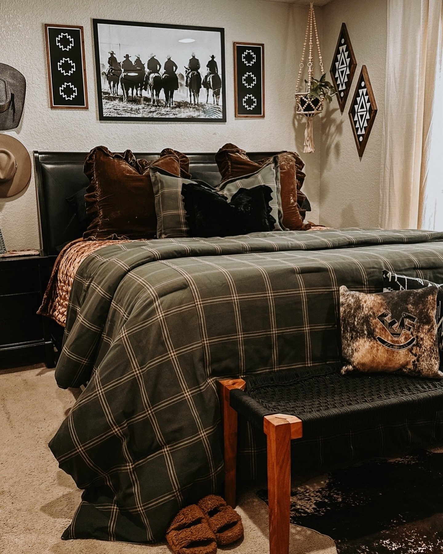 A Western bedroom featuring a black upholstered headboard, a dark olive green plaid duvet, velvet pillows, and a framed black and white photo of cowboys on horseback.