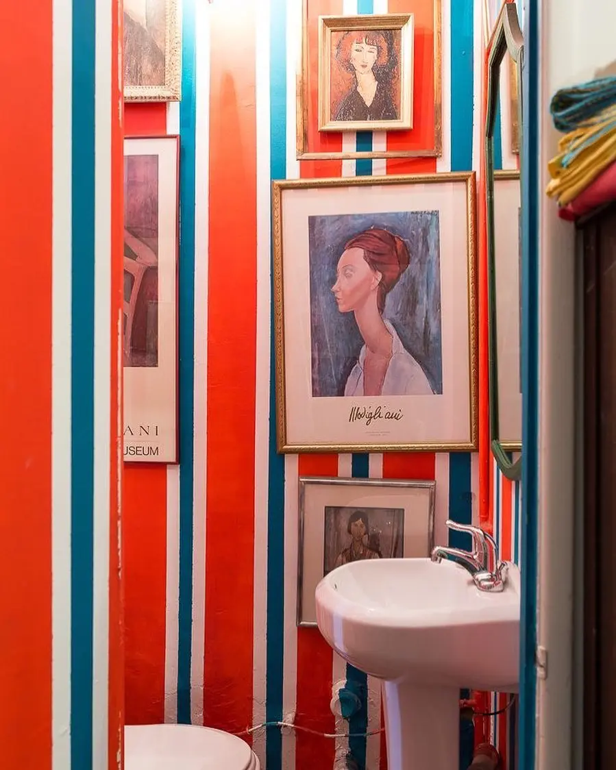 A small, maximalist powder room with bright red, white, and blue striped walls, featuring a gallery wall of framed portraits and prints.