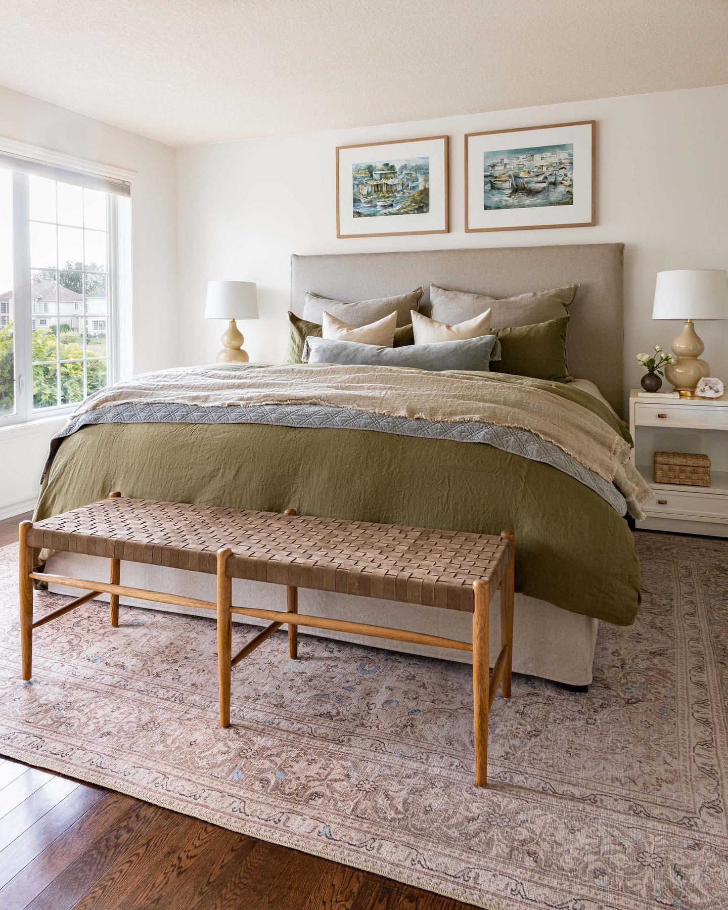 A bright bedroom featuring a bed with an olive green duvet, a woven leather bench, and a vintage-style patterned rug.