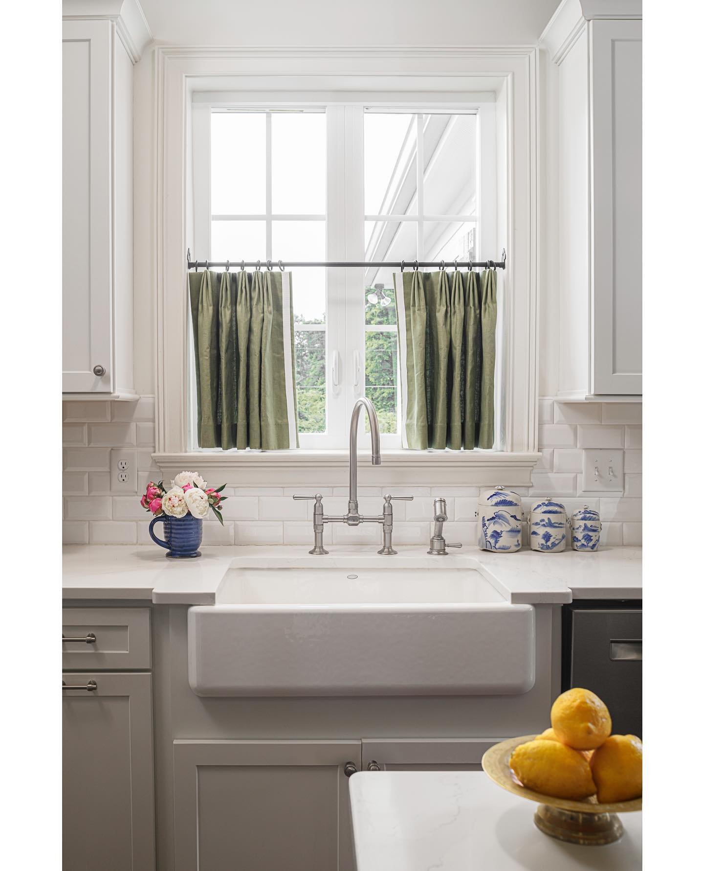 White kitchen with a farmhouse sink and subway tile, featuring two short, deep sage green pleated cafe curtains hung on a black rod.