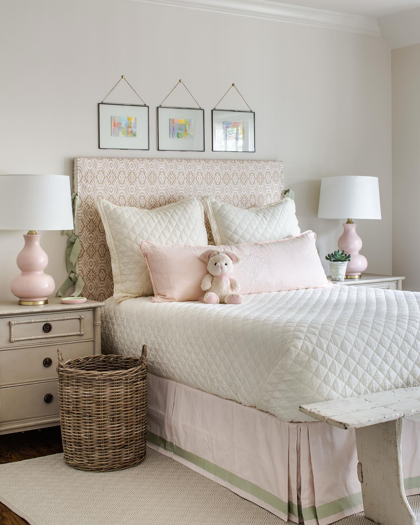 Subtle pink bedroom with an antique white nightstand, patterned fabric headboard, pink gourd lamps, and a quilted white comforter.