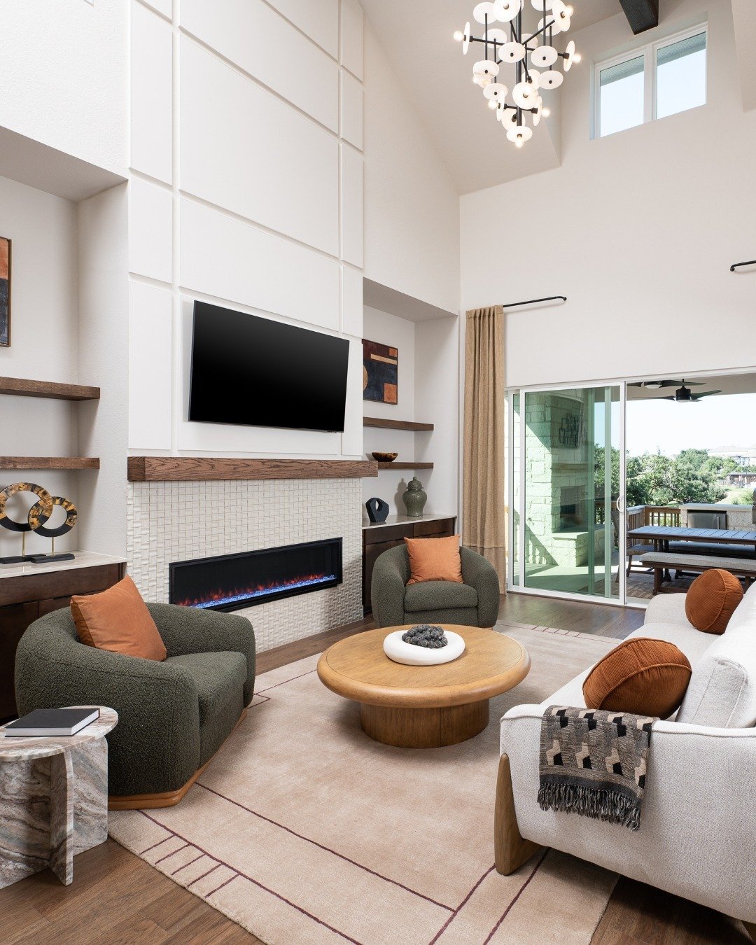 Luxurious two-story living room with a feature wall of white geometric paneling, a linear electric fireplace with a white mosaic tile surround, a wood mantel, and flanking wood shelves.