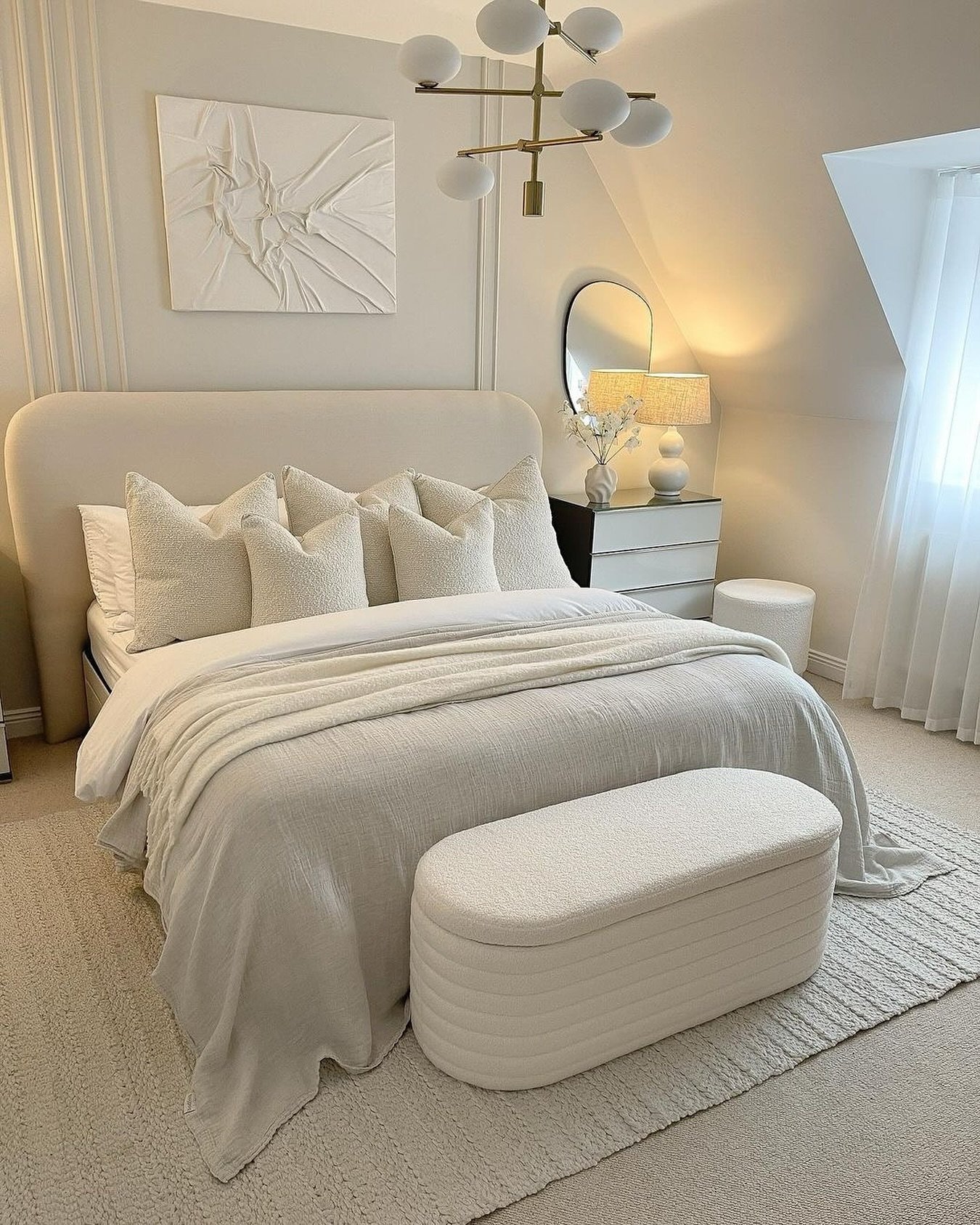 Attik-style bedroom featuring an oversized rounded headboard, a white horizontal ribbed ottoman, and a modern brass and white globe chandelier.