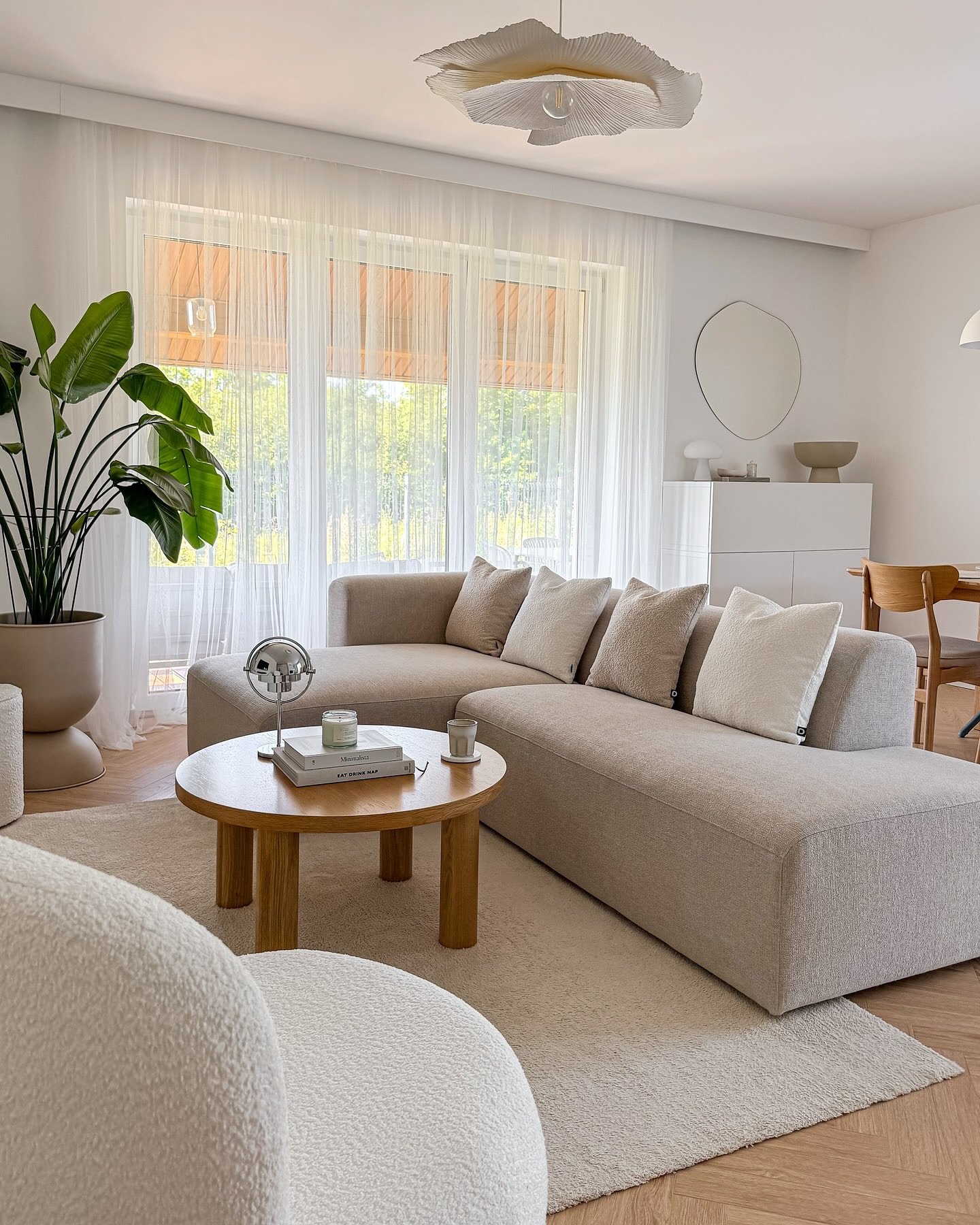 A bright neutral living room with a low, curved beige sectional sofa, a round wood coffee table, and a large plant next to the window.