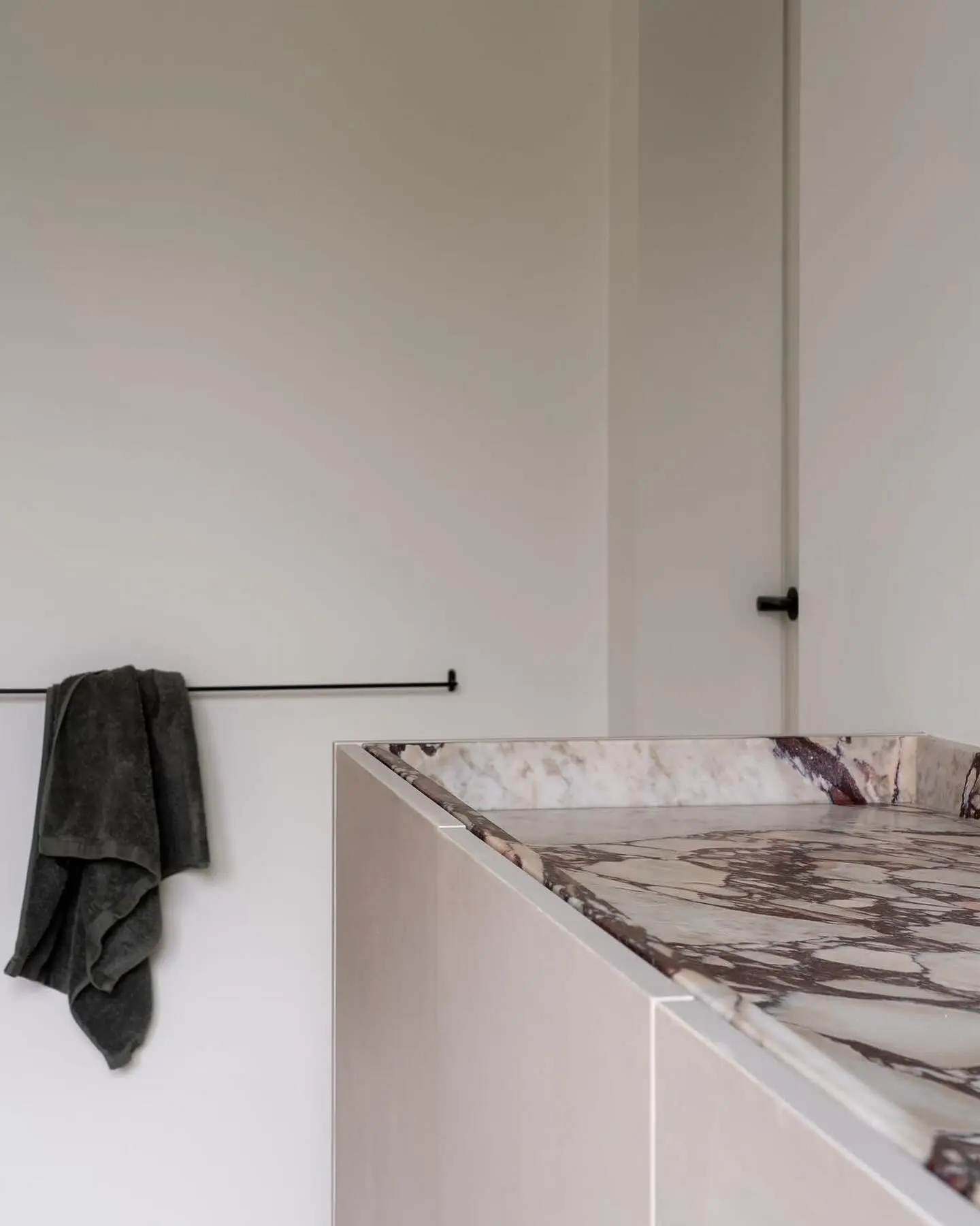 A single, sleek matte black towel bar holding a dark green bath towel, mounted low on a plain white wall above a vanity with a patterned marble countertop.