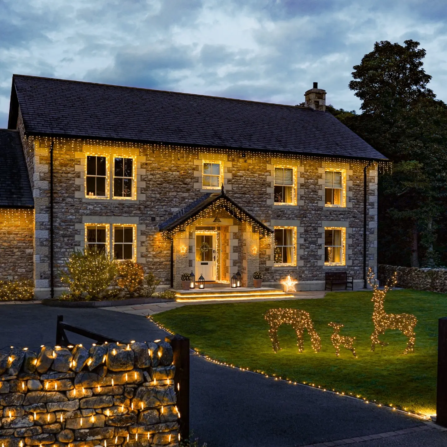 A stone house is decorated with warm white icicle lights across the rooflines and windows, with string lights outlining the driveway and a stone wall, featuring light-up reindeer figures on the lawn.