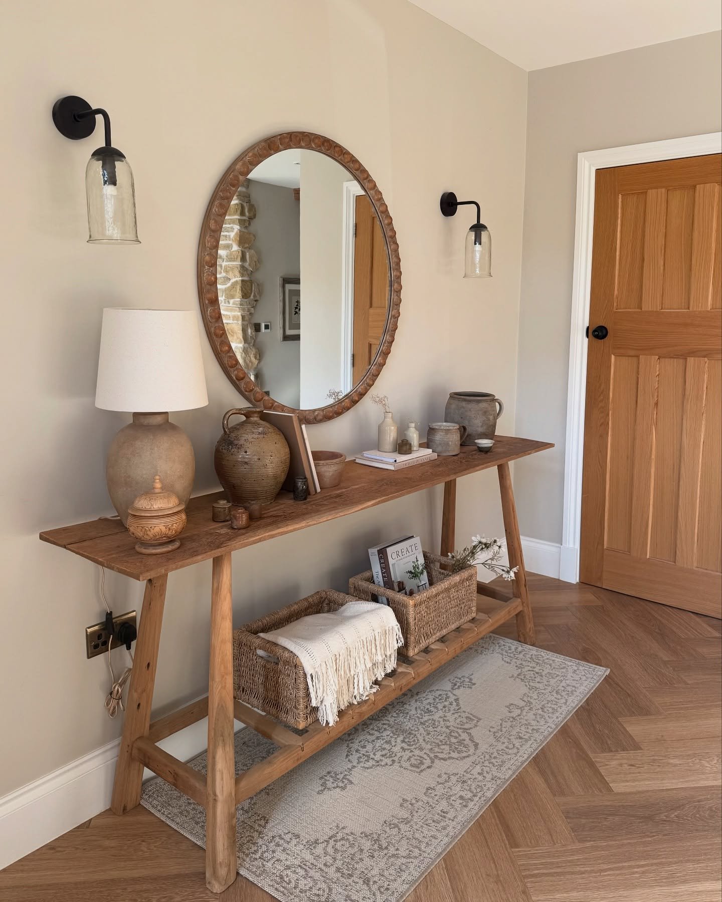 A rustic wood console table in a light taupe hallway with a large wooden-framed round mirror and an array of textured pottery and lighting.