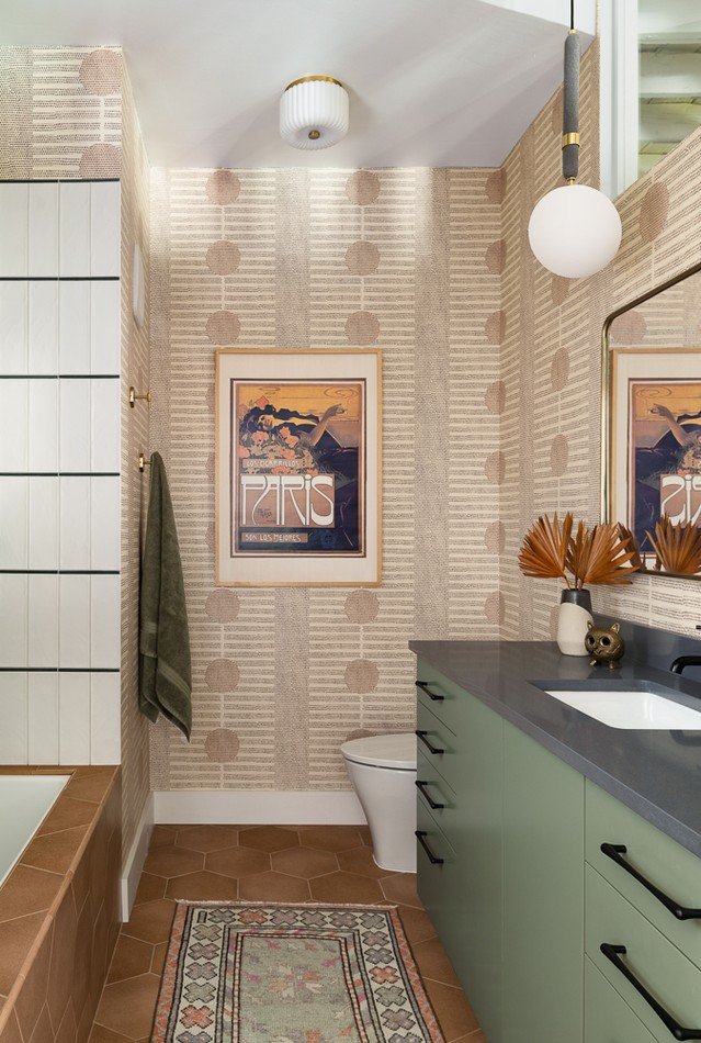 A small, eclectic bathroom featuring patterned beige and pink wallpaper, a sage green vanity with a dark countertop, terracotta hexagonal floor tiles, and a small, patterned runner rug in muted pinks and greens.