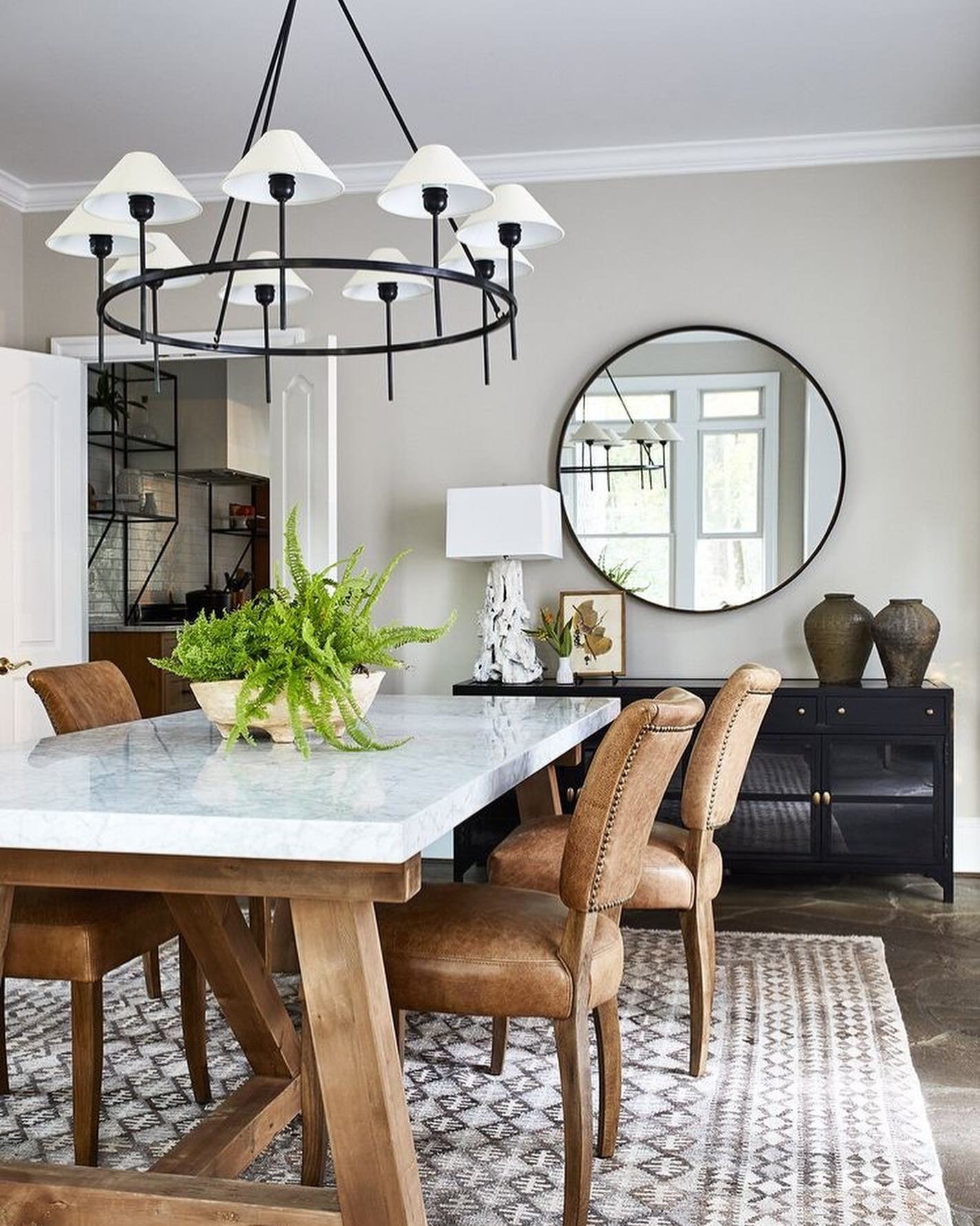 A dining room with a white marble-top table on a rustic wooden base, warm brown leather studded chairs, and a large black metal ring chandelier.