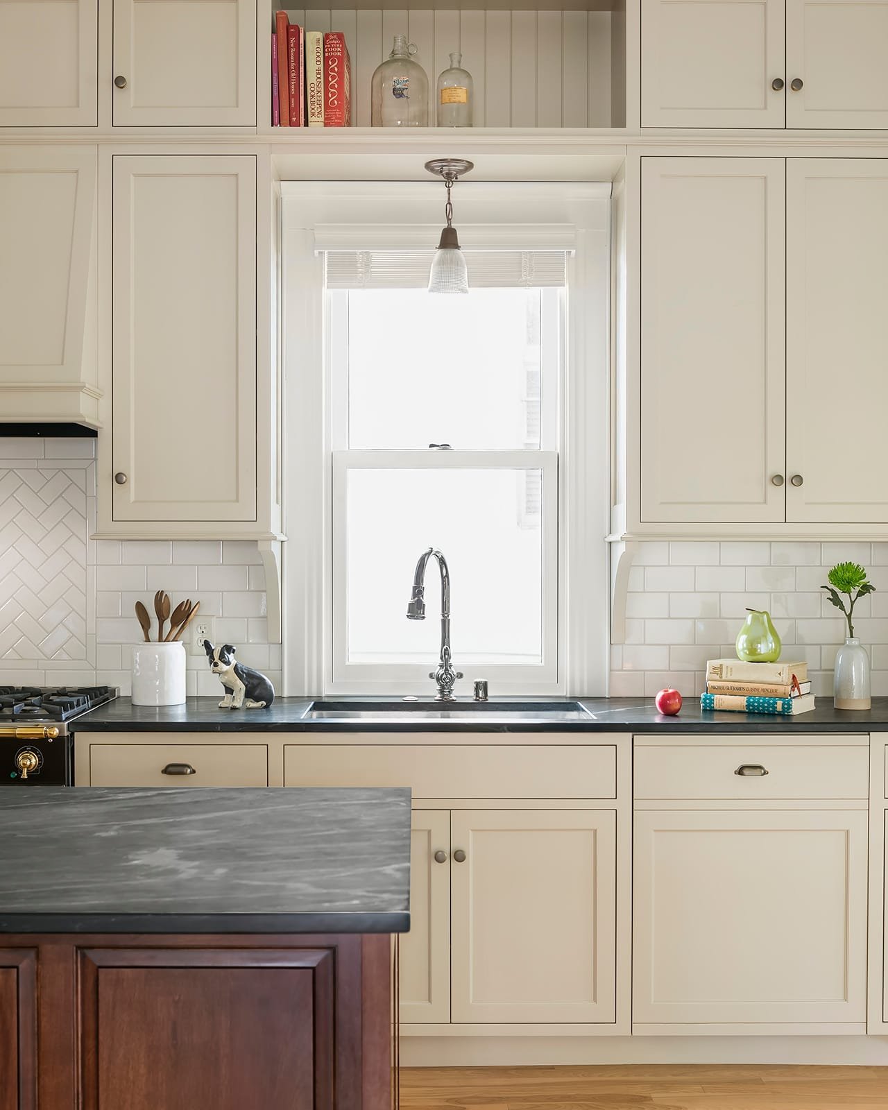 Kitchen with creamy white cabinetry and dark countertops. A bright single sash window over the sink is completely framed by cabinets, including a shallow shelf above it.