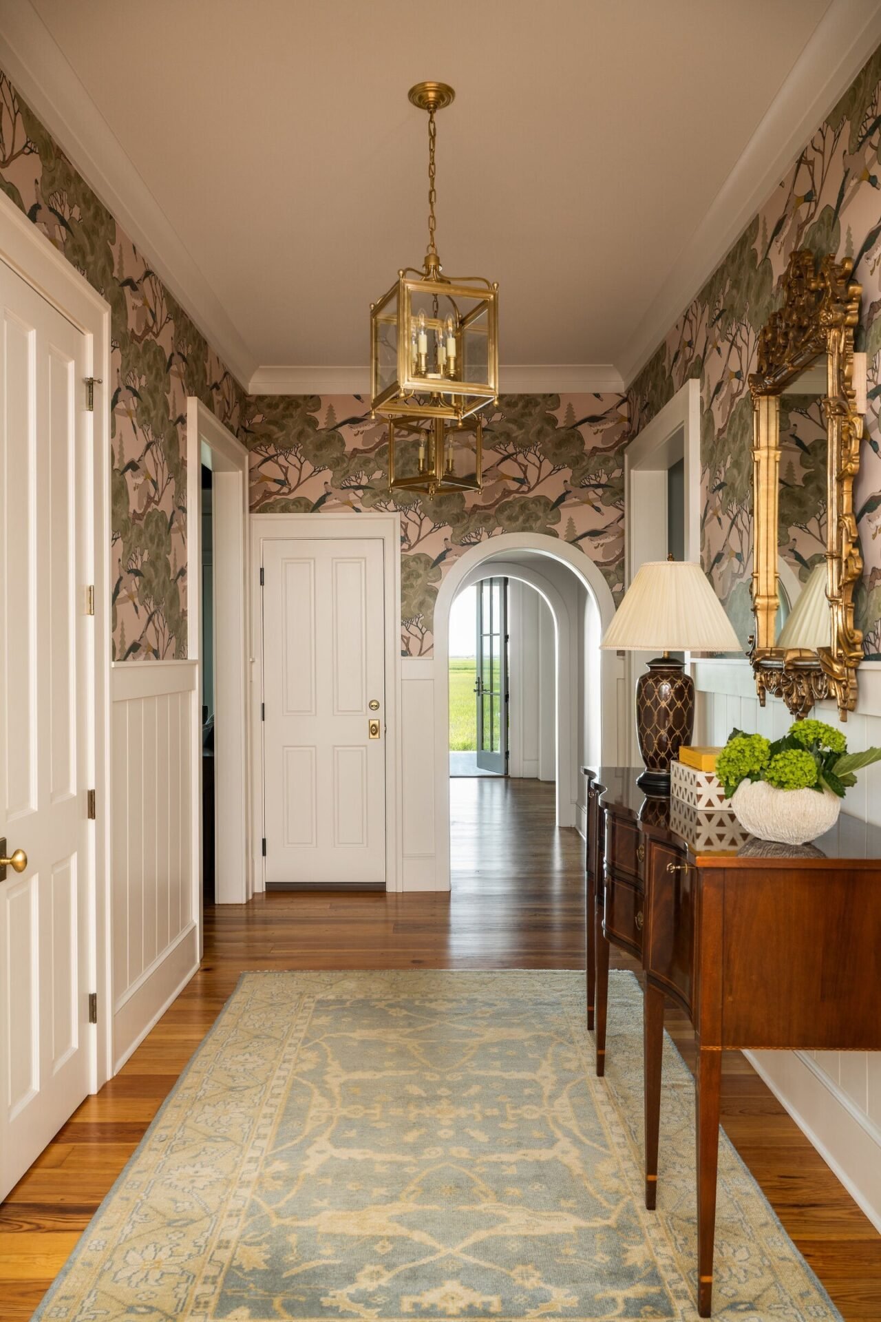 Traditional hallway with unique pink and green patterned wallpaper above white wainscoting, wood floor, blue and gold runner rug, and a central brass lantern pendant.
