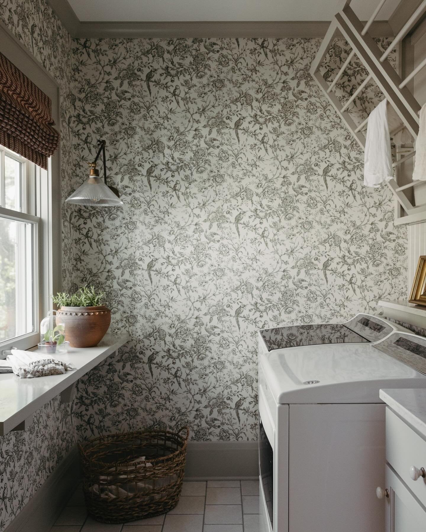 A small laundry room decorated with a detailed black and white toile or botanical wallpaper and featuring a white floating shelf beneath a window and a large fold-down wooden drying rack.