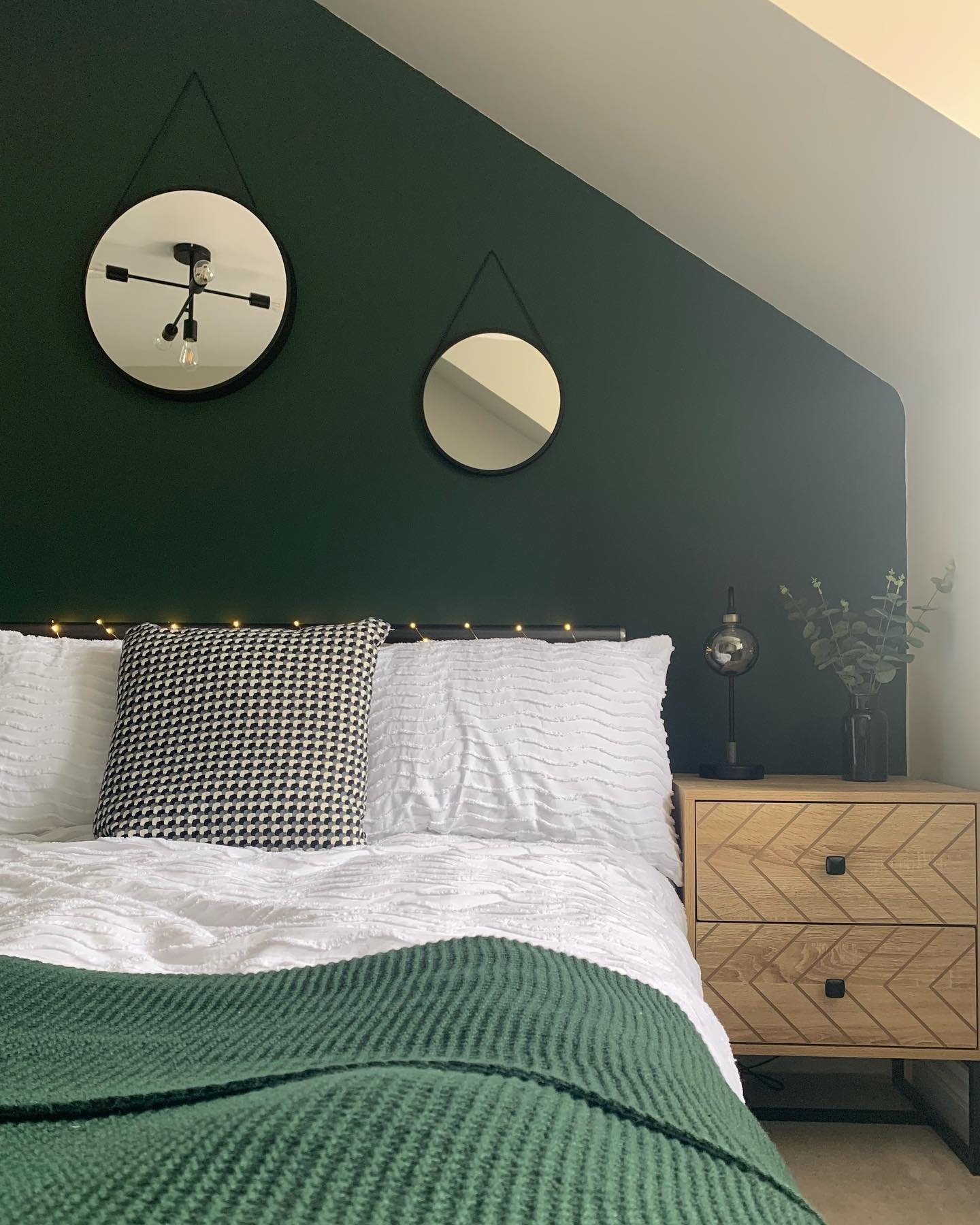 Bedroom featuring a sloped accent wall painted deep green, with chevron-patterned wooden nightstands and hanging circular mirrors.