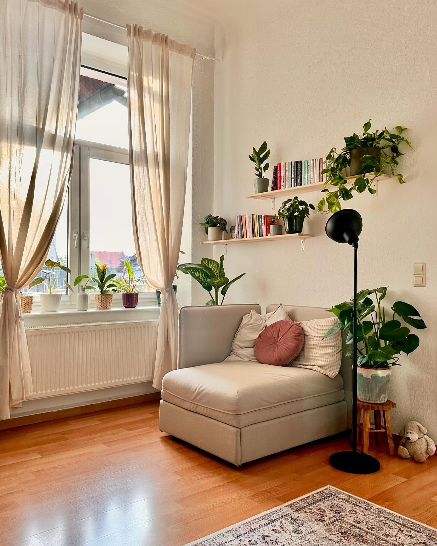 Modern reading nook with a neutral daybed, light wood floating shelves with books and plants, and a black floor lamp next to a large window.