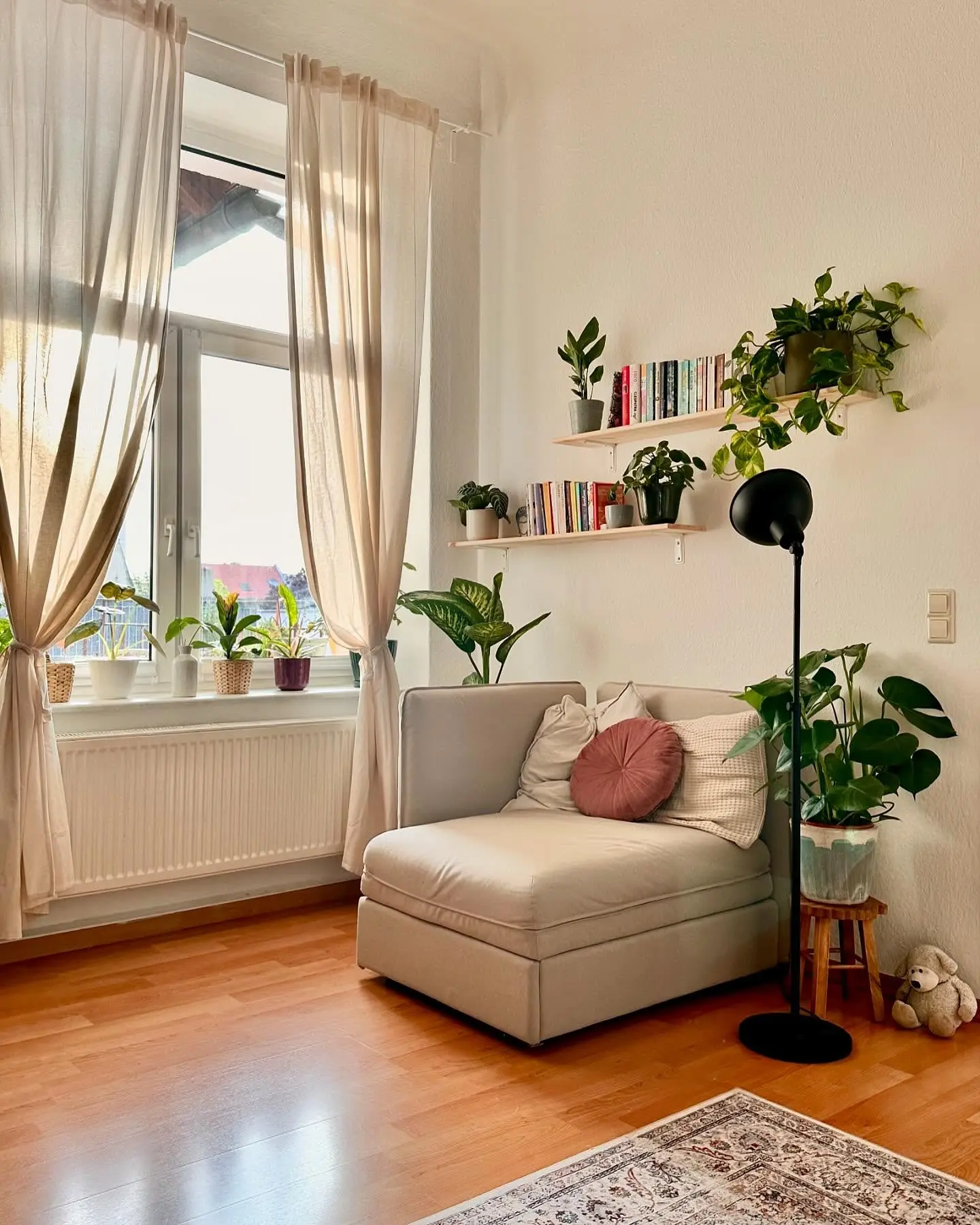 Modern reading nook with a neutral daybed, light wood floating shelves with books and plants, and a black floor lamp next to a large window.
