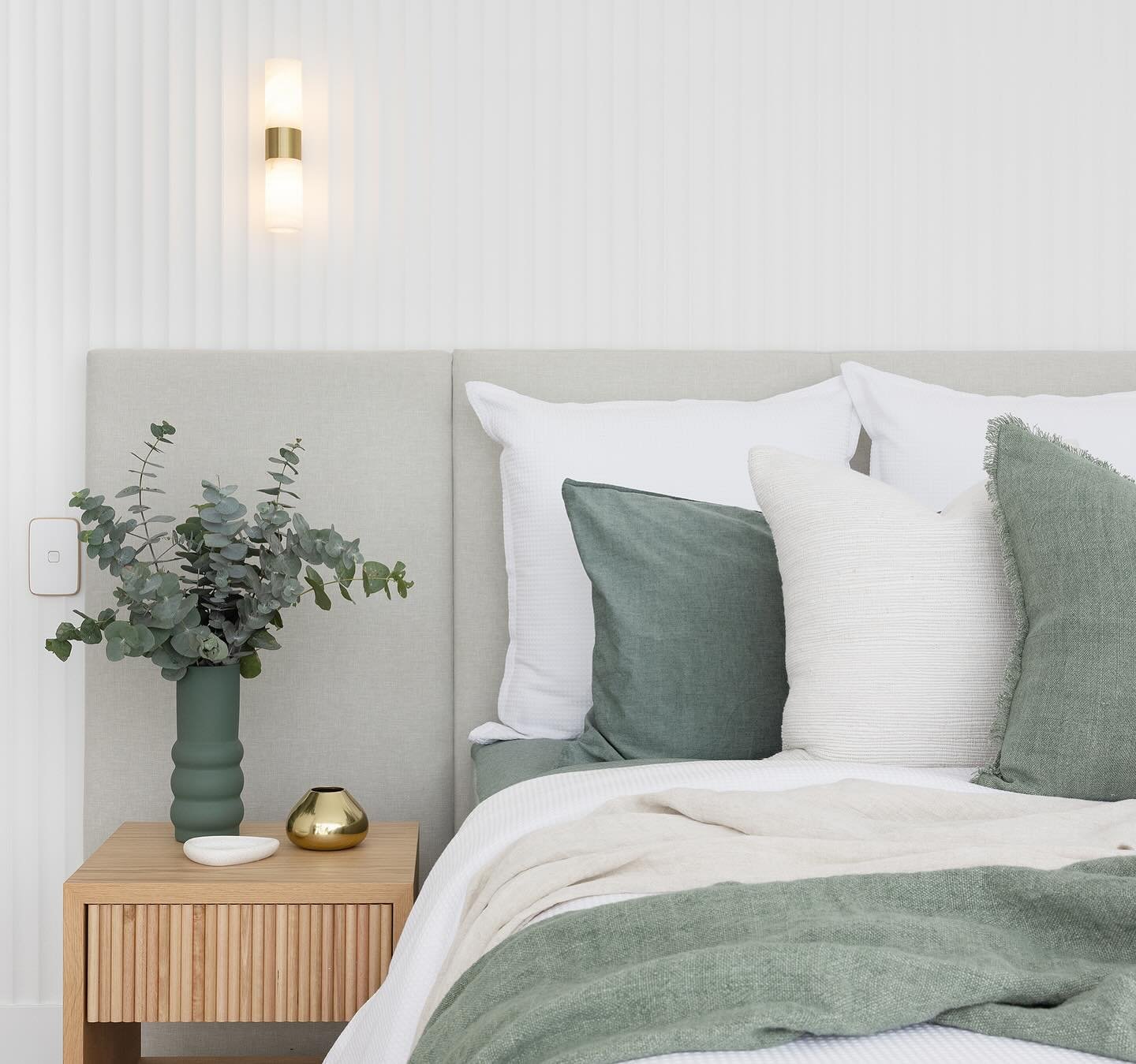 Bedroom detail showing a light oak bedside table, eucalyptus in a green vase, and sage green and white pillows against a fluted white wall.
