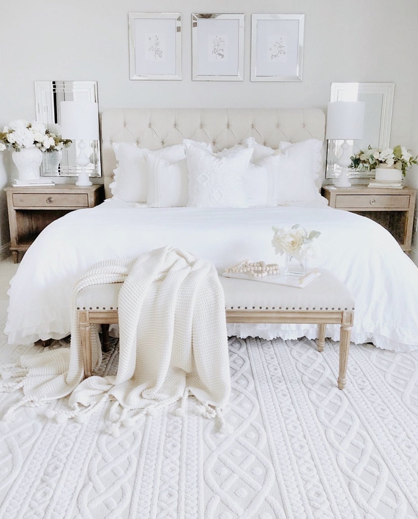 A bright, all-white bedroom with a tufted headboard, white bedding, a thick cable-knit rug, and a small upholstered bench with nailhead trim at the foot of the bed.