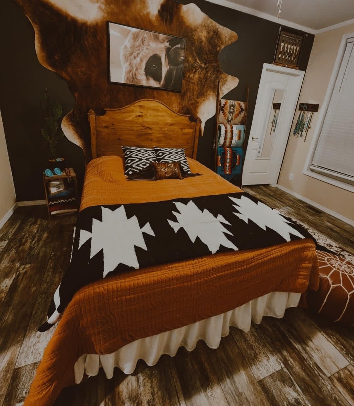 A Western bedroom with a black accent wall, a natural wood headboard, a large cowhide layered on the wall, and bright orange bedding.