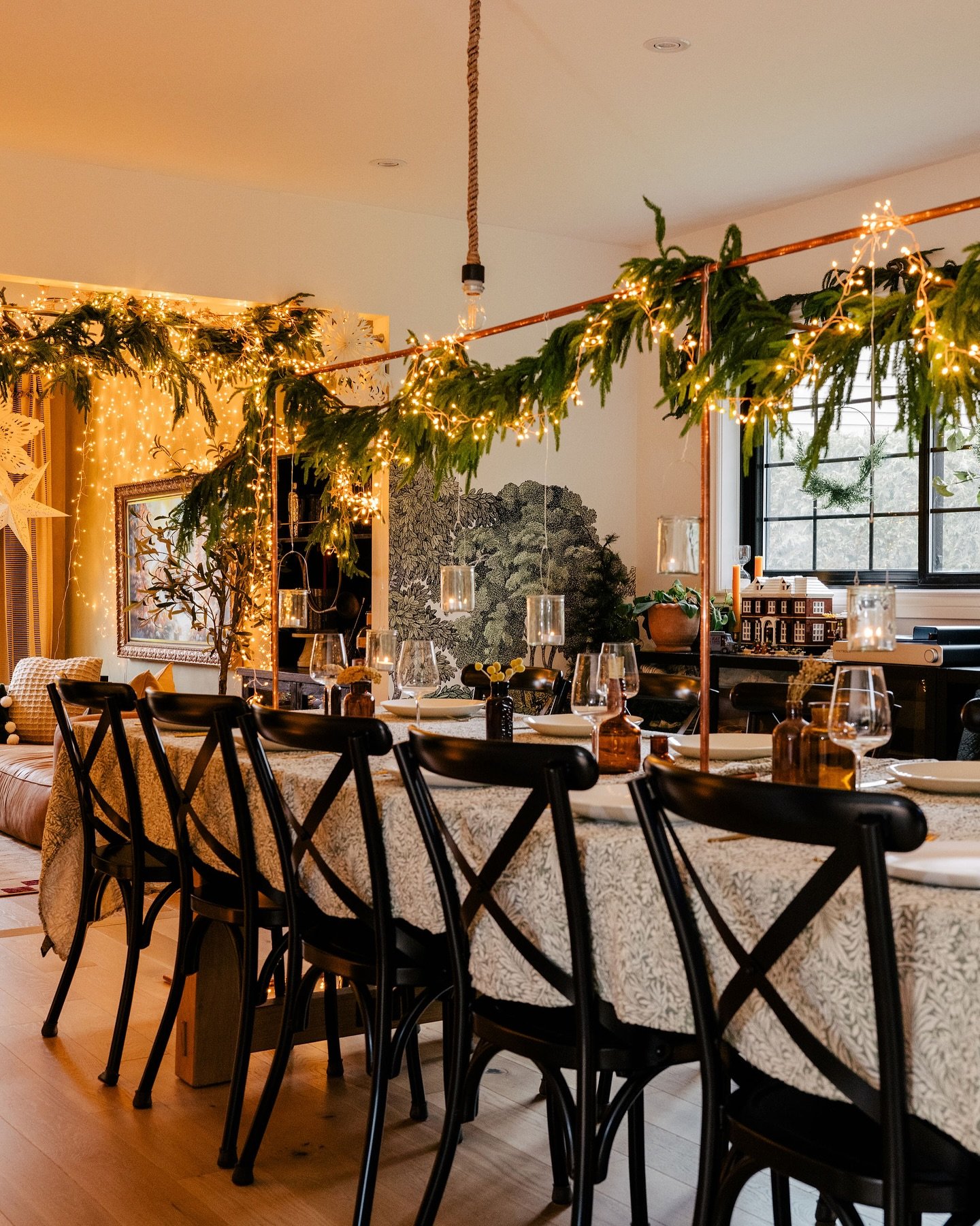 A dining table covered in a patterned white tablecloth, set with dark amber glasses, and featuring an overhead structure made of copper pipes wrapped in evergreen garland and tiny string lights.