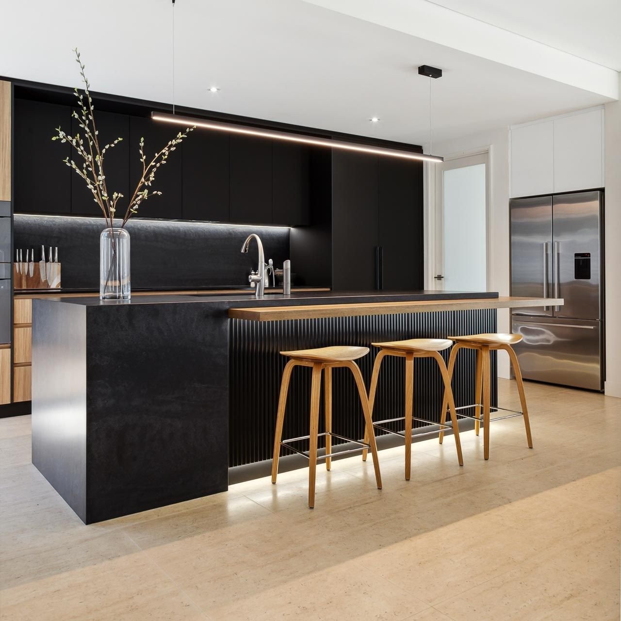 Ultra-modern kitchen featuring matte black cabinetry, light wood accents, a black kitchen island with a fluted wood section, and a linear LED pendant light.